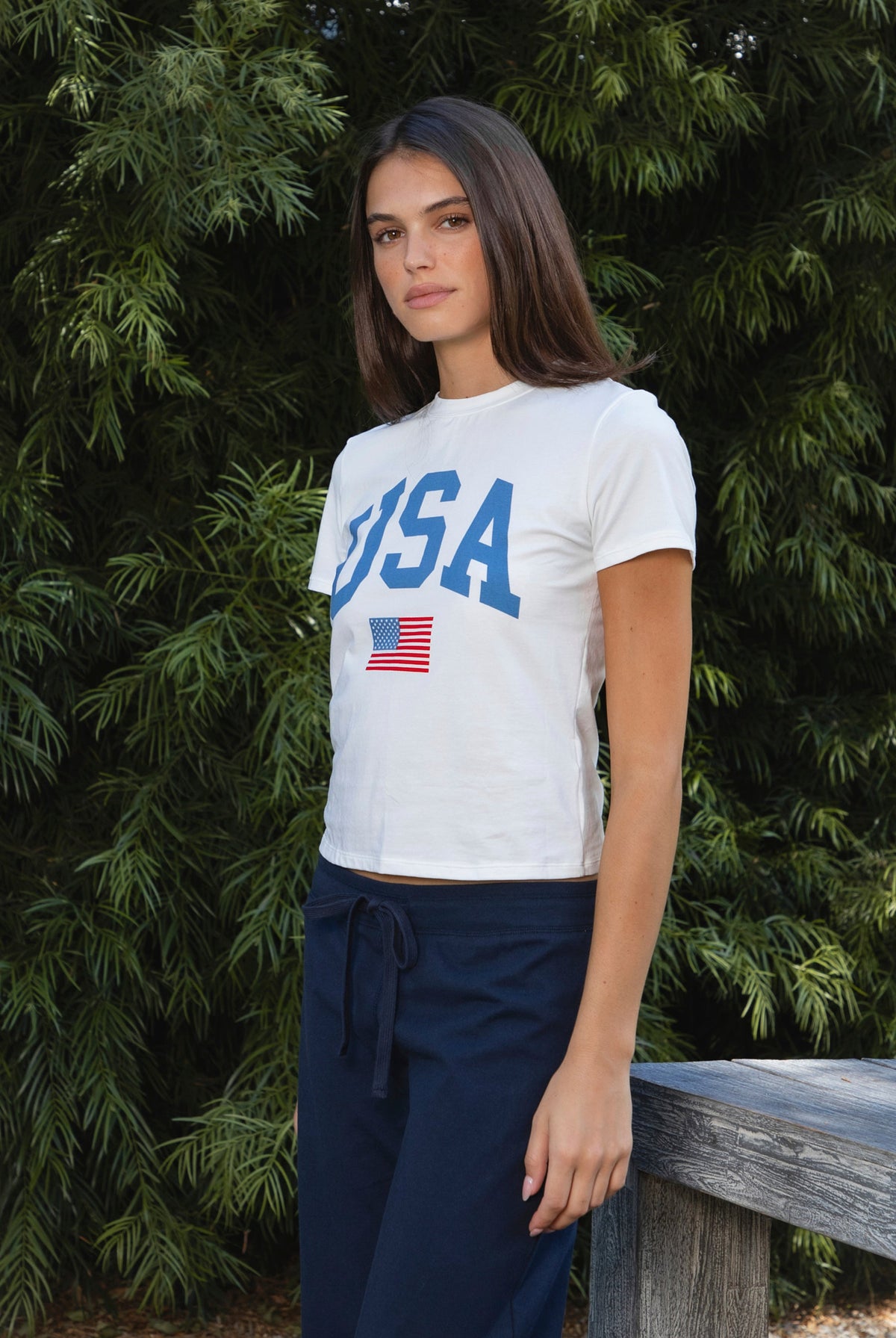 Woman wearing LIBERTY classic USA graphic crewneck baby tee featuring soft single jersey cotton stretch fabric, fitted short sleeve baby tee silhouette, clean cropped length, classic crew neckline, bold USA graphic across the front, American flag graphic detail below, and straight fitted hemline.