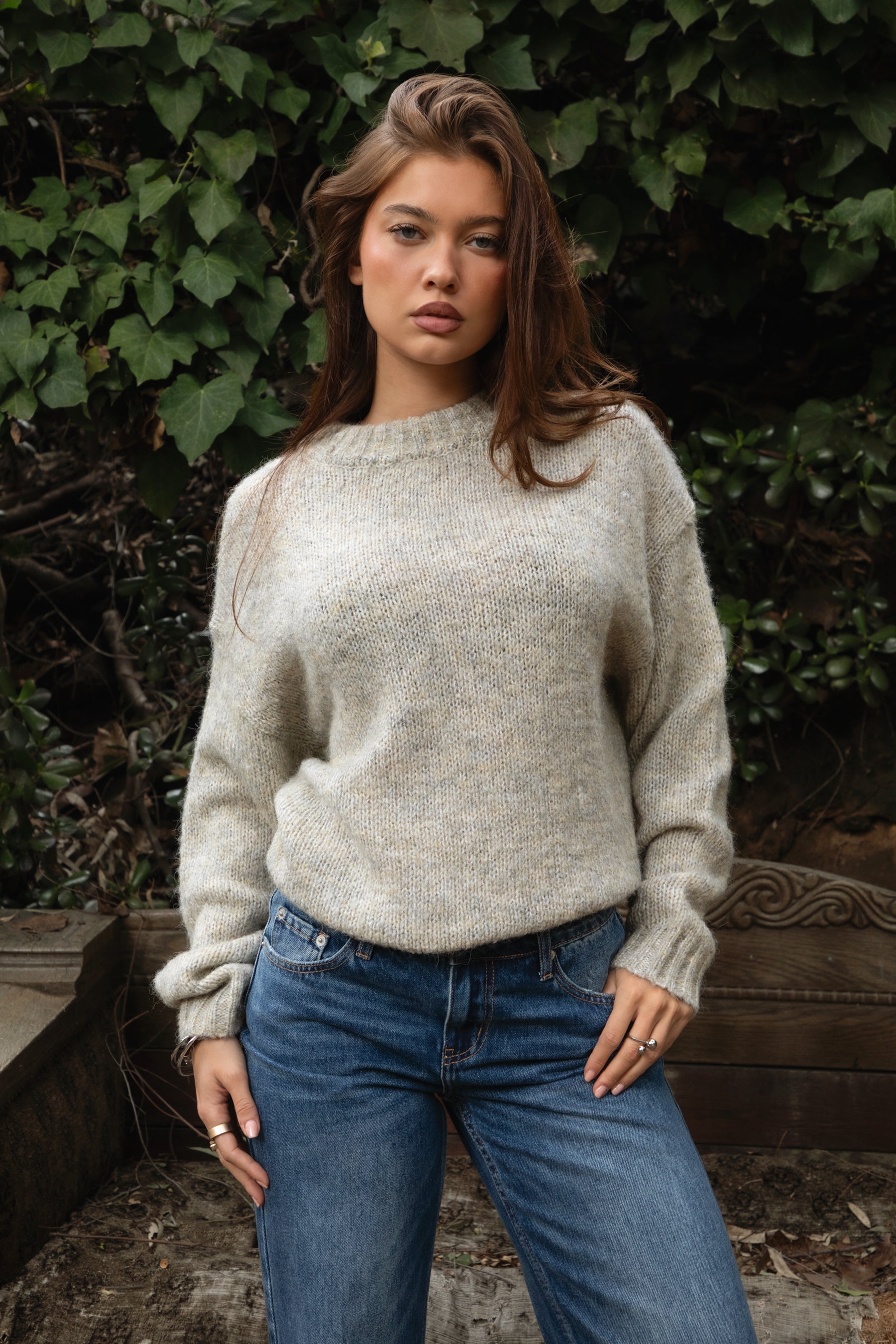 Woman wearing TIC TOC TF31840 SELYNA heather oatmeal knit pullover sweater with a soft wool-blend texture, relaxed oversized fit, drop shoulders, and long ribbed-cuff sleeves paired with medium-wash denim jeans in a cozy outdoor garden setting.