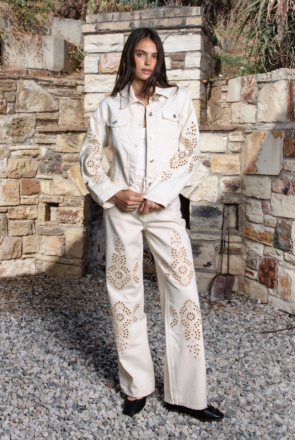 Woman wearing ALBA embroidered wide leg denim pants featuring structured cotton blend denim, high rise waist, classic four pocket design, intricate floral eyelet embroidery throughout the legs, relaxed wide leg silhouette, full length fit, button and zip fly closure, and belt loops along the waistline.