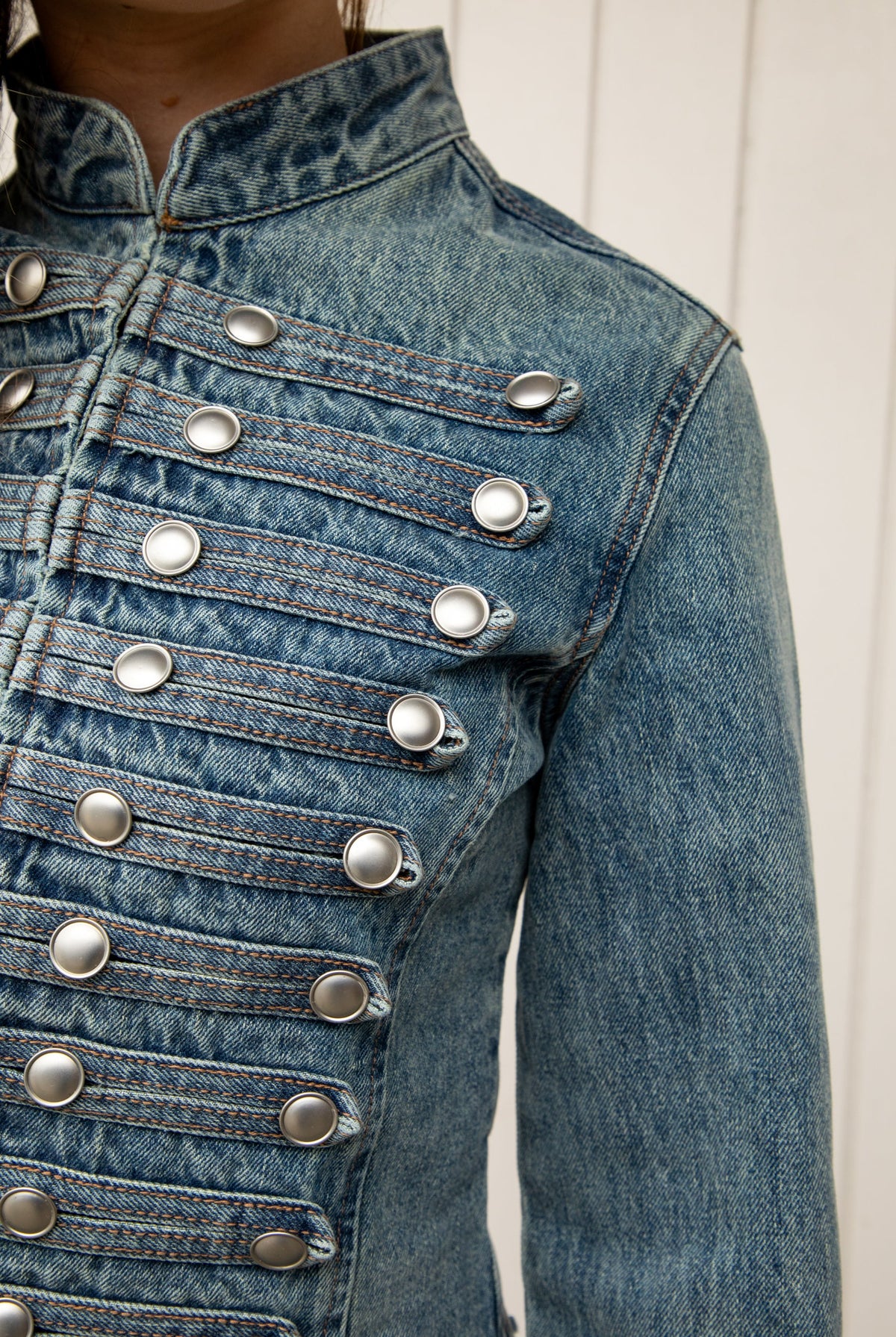 Woman wearing REMINGTON military semi crop denim jacket featuring cotton blend denim, structured tailored fit, military inspired horizontal strap panels, matte metal button accents, stand collar, hidden hook and eye front closure, long sleeves with metal button cuffs, and cropped hemline.