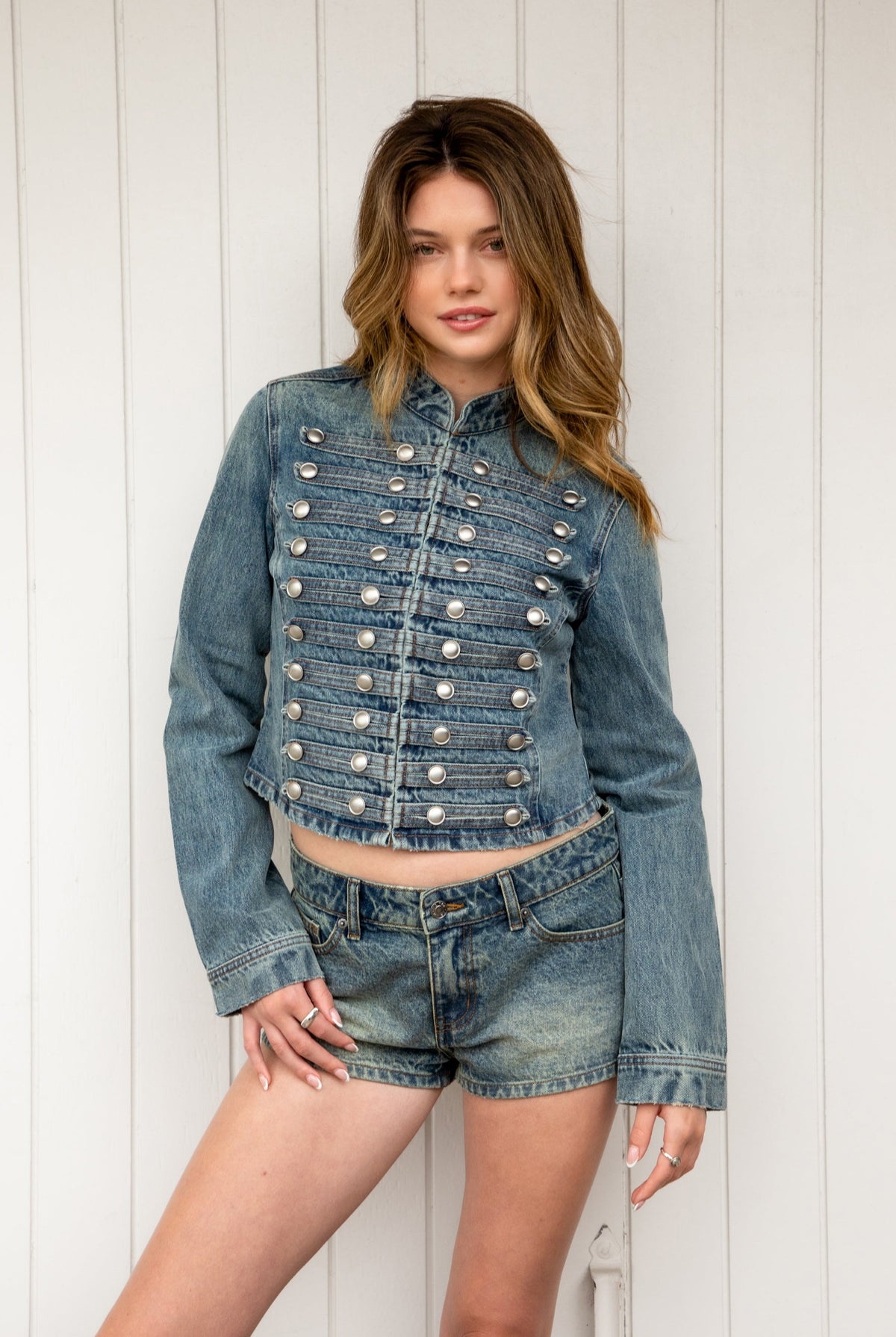 Woman wearing REMINGTON military semi crop denim jacket featuring cotton blend denim, structured tailored fit, military inspired horizontal strap panels, matte metal button accents, stand collar, hidden hook and eye front closure, long sleeves with metal button cuffs, and cropped hemline.
