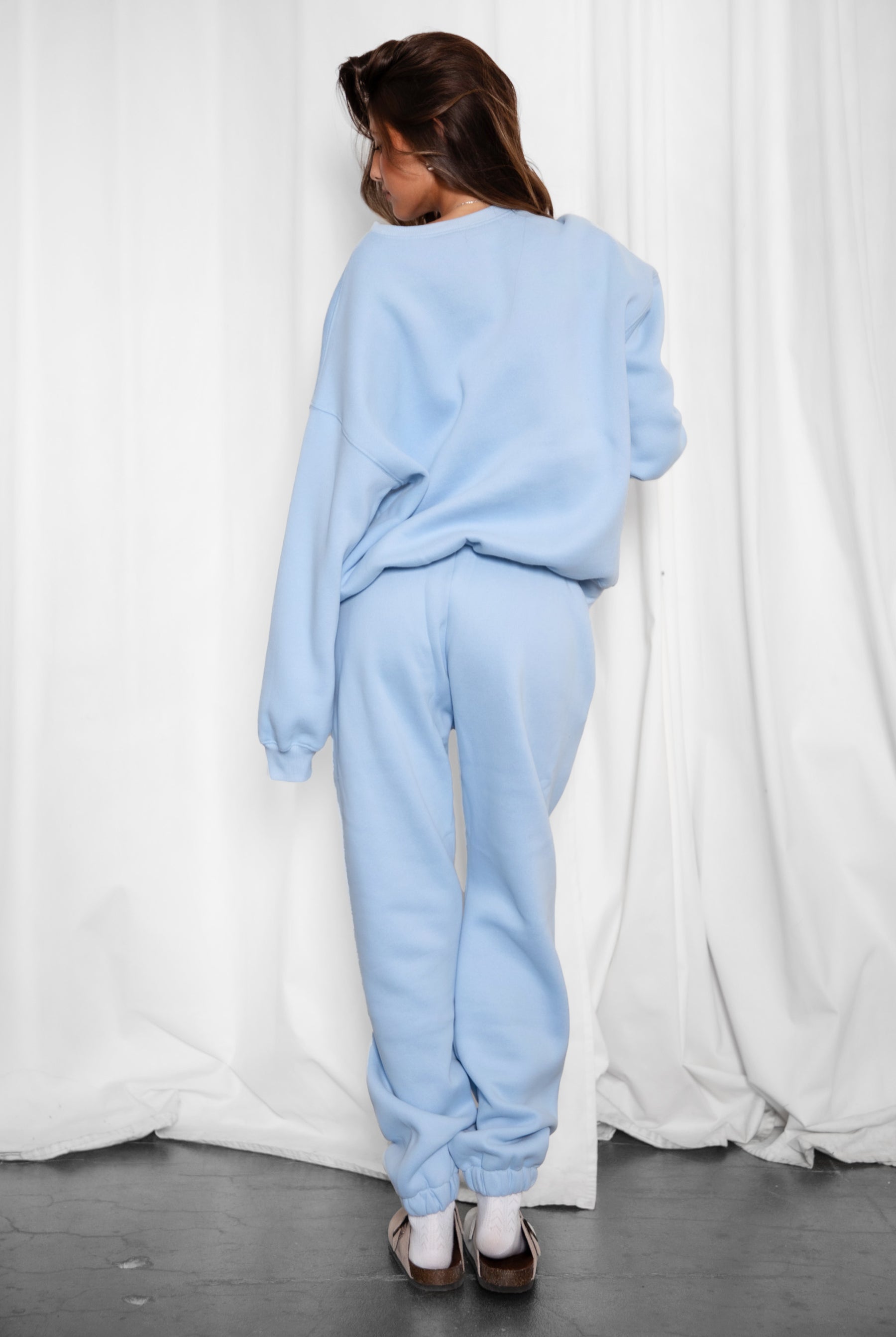Person wearing a light blue tracksuit standing against a white curtain.