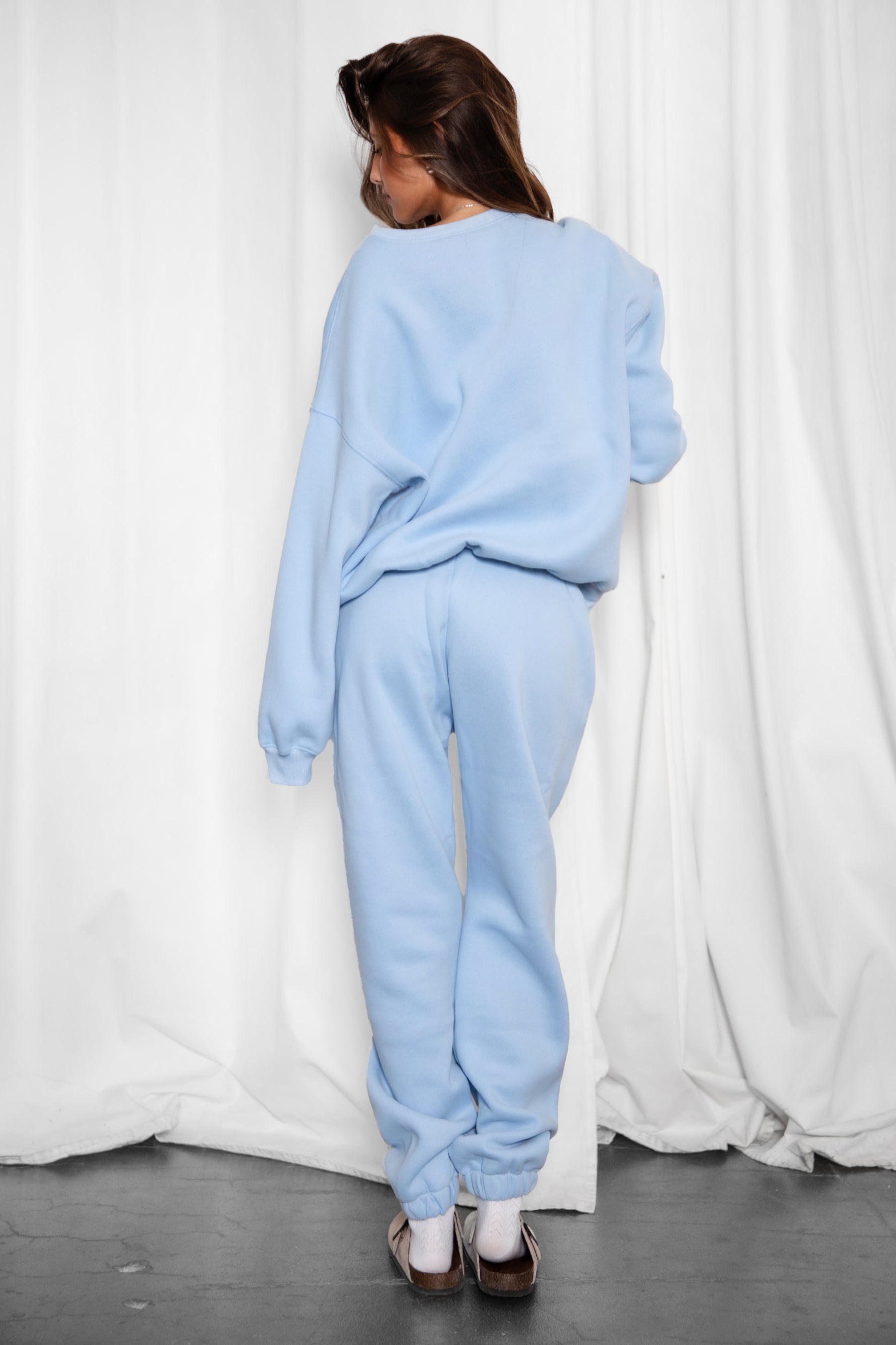 Person wearing a light blue tracksuit standing against a white curtain.