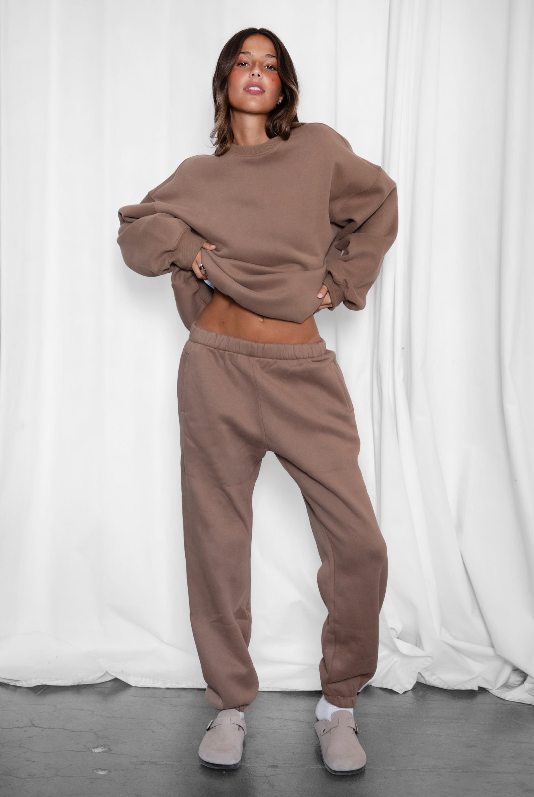 Person wearing a brown sweatshirt and pants set against a white curtain background