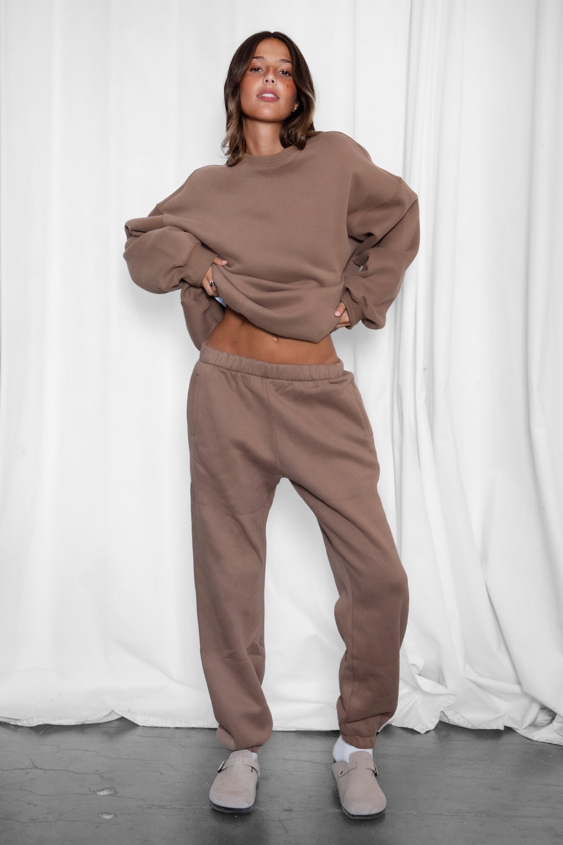 Person wearing a brown sweatshirt and pants set against a white curtain background