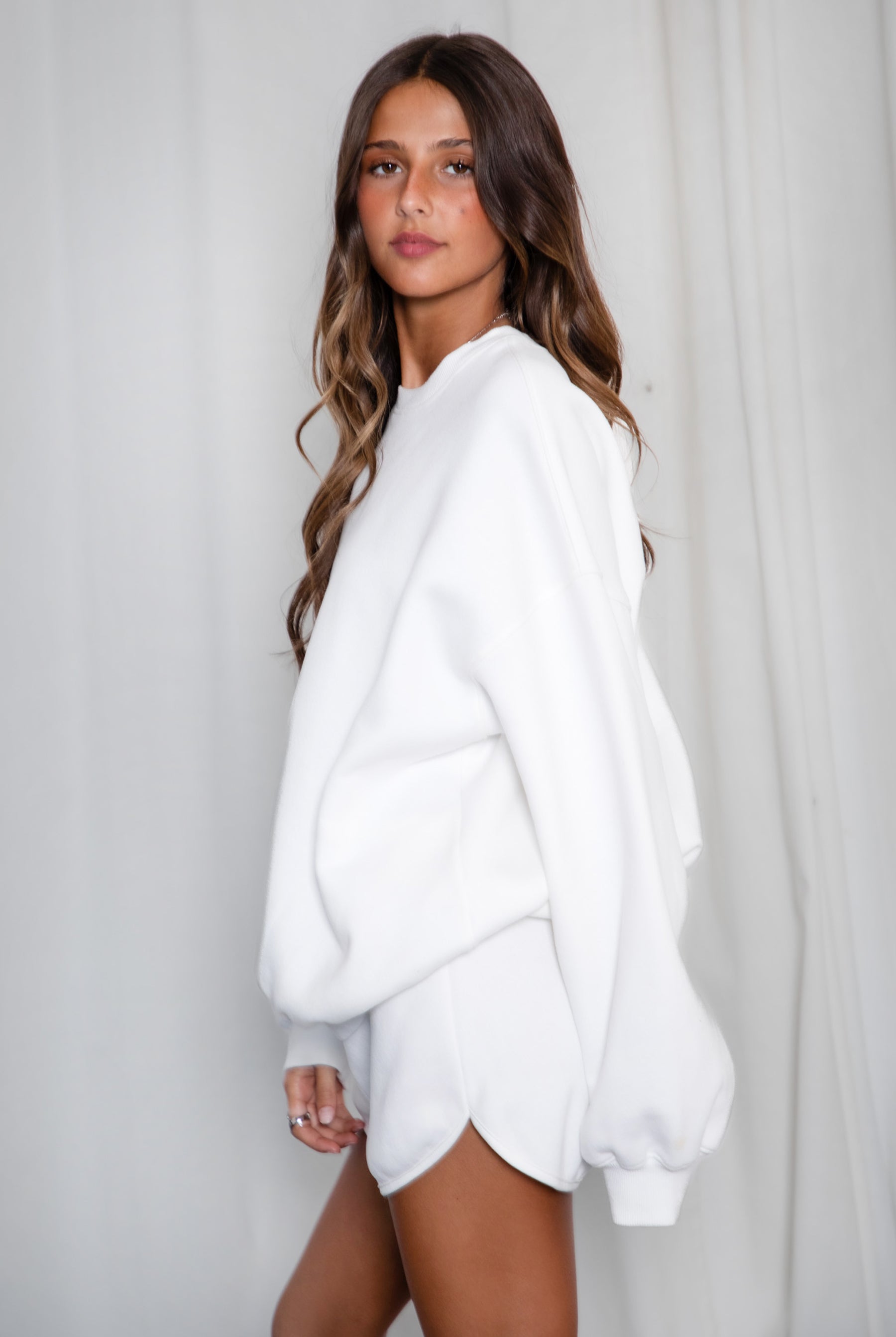 Woman wearing a white oversized sweater and shorts against a plain background