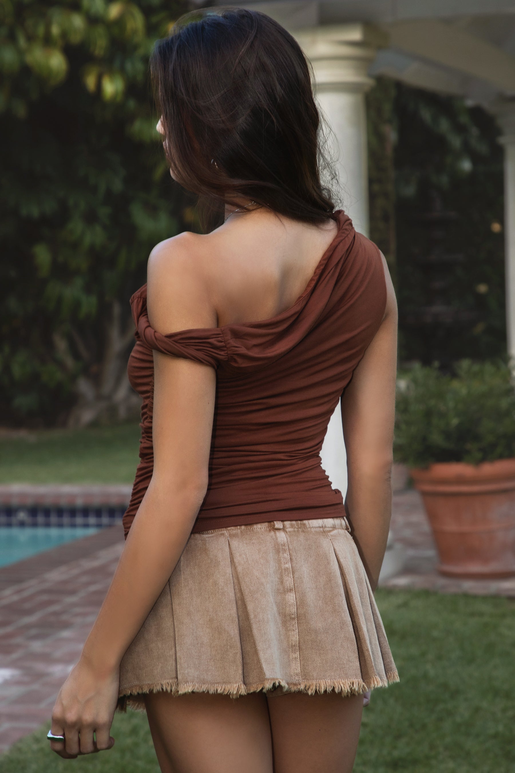 Women wearing TIC TOC SHANTAL One Shoulder Top in copper with twisted strap, ruched seams, and studded torso detail paired with a denim brown pleated mini skirt.