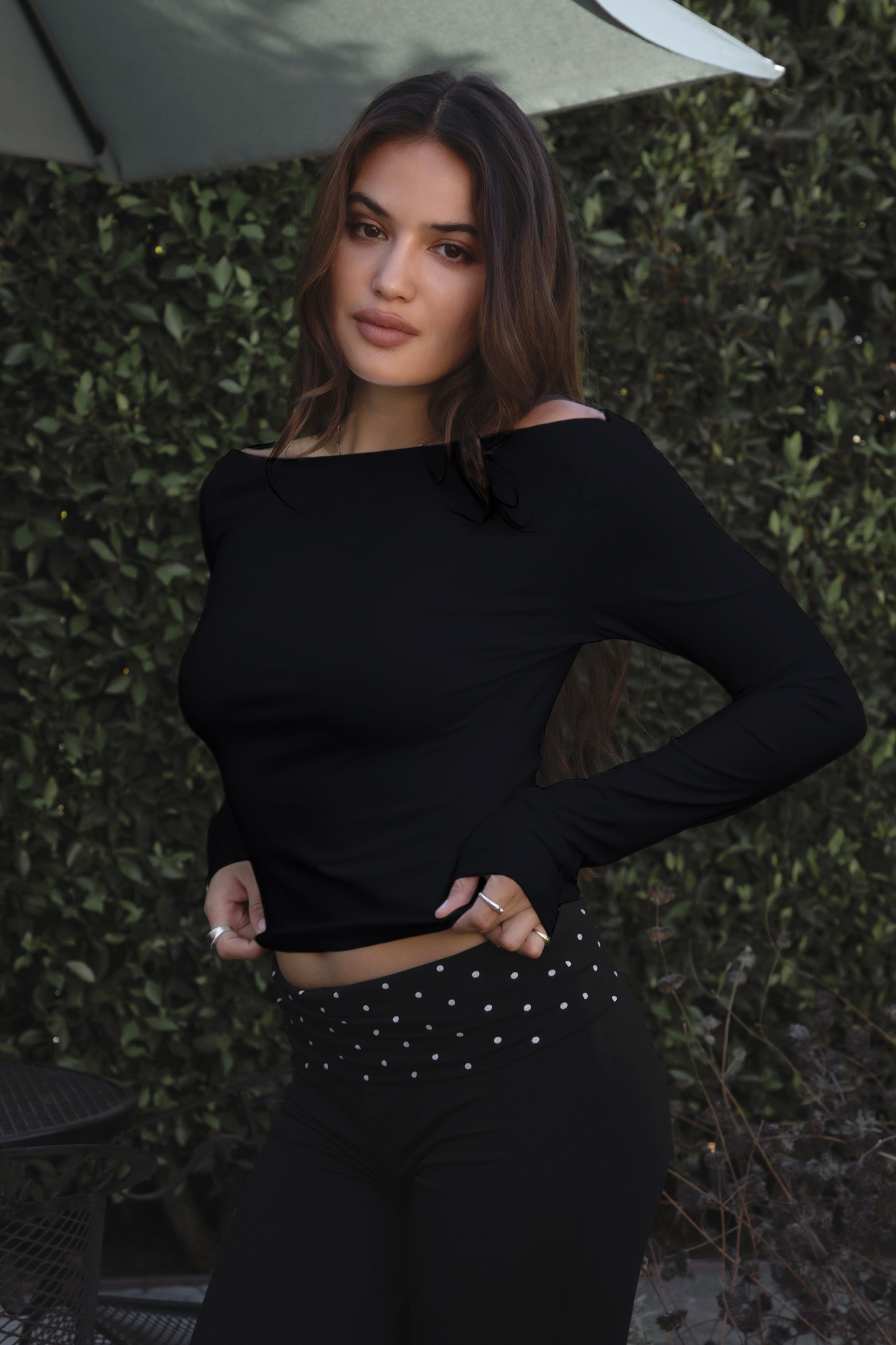 Model wearing TIC TOC TH31573BA RAYAE asymmetric off-the-shoulder jersey top with wide neckline and draped long sleeves paired with polka dot black capri pants in an outdoor setting.