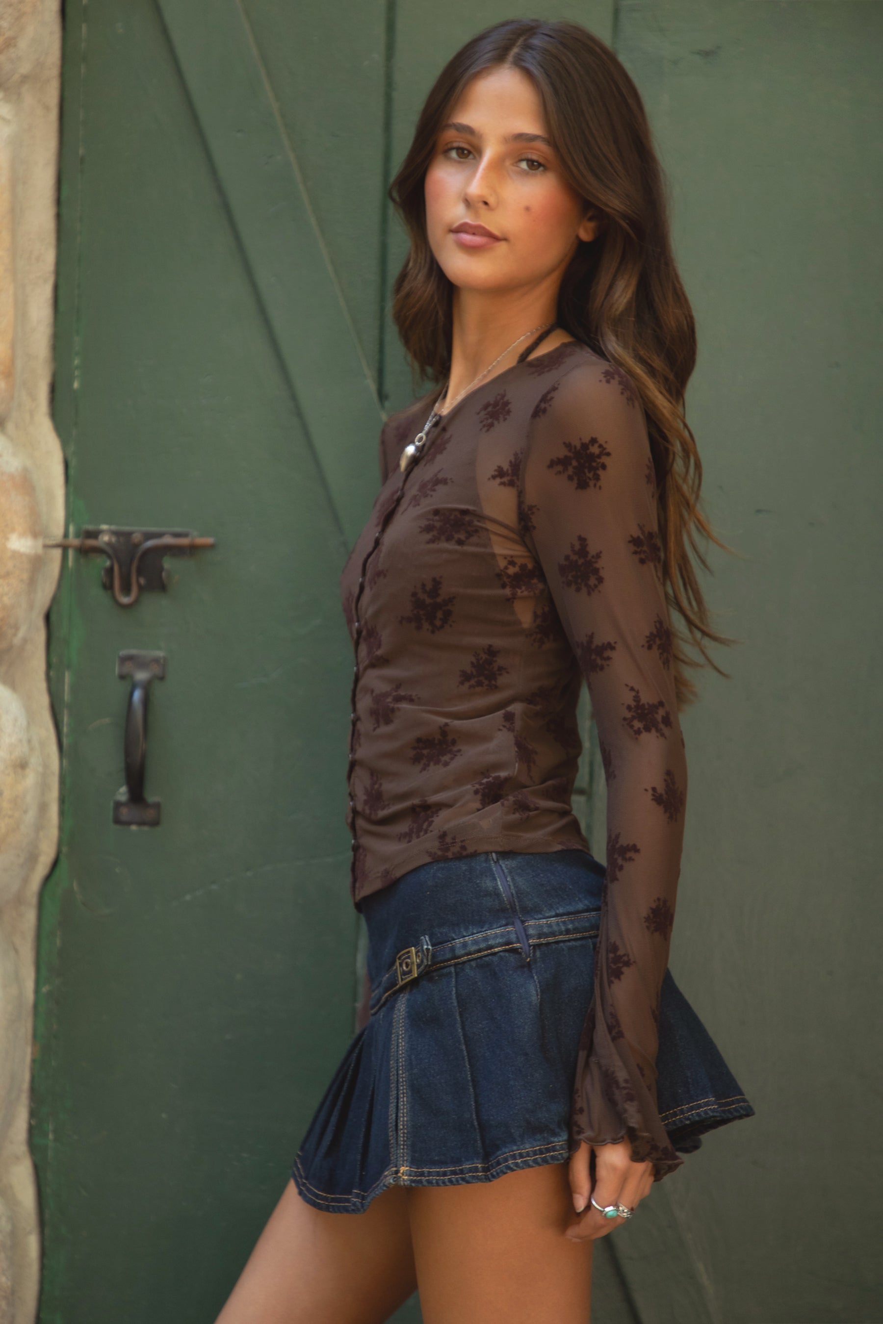 Woman wearing TIC TOC LINETTE Floral Flocked Mesh Long Sleeve Top in brown with high mock neckline, bell sleeves, and button front detail and blue denim skirt standing against a stone wall and green door.