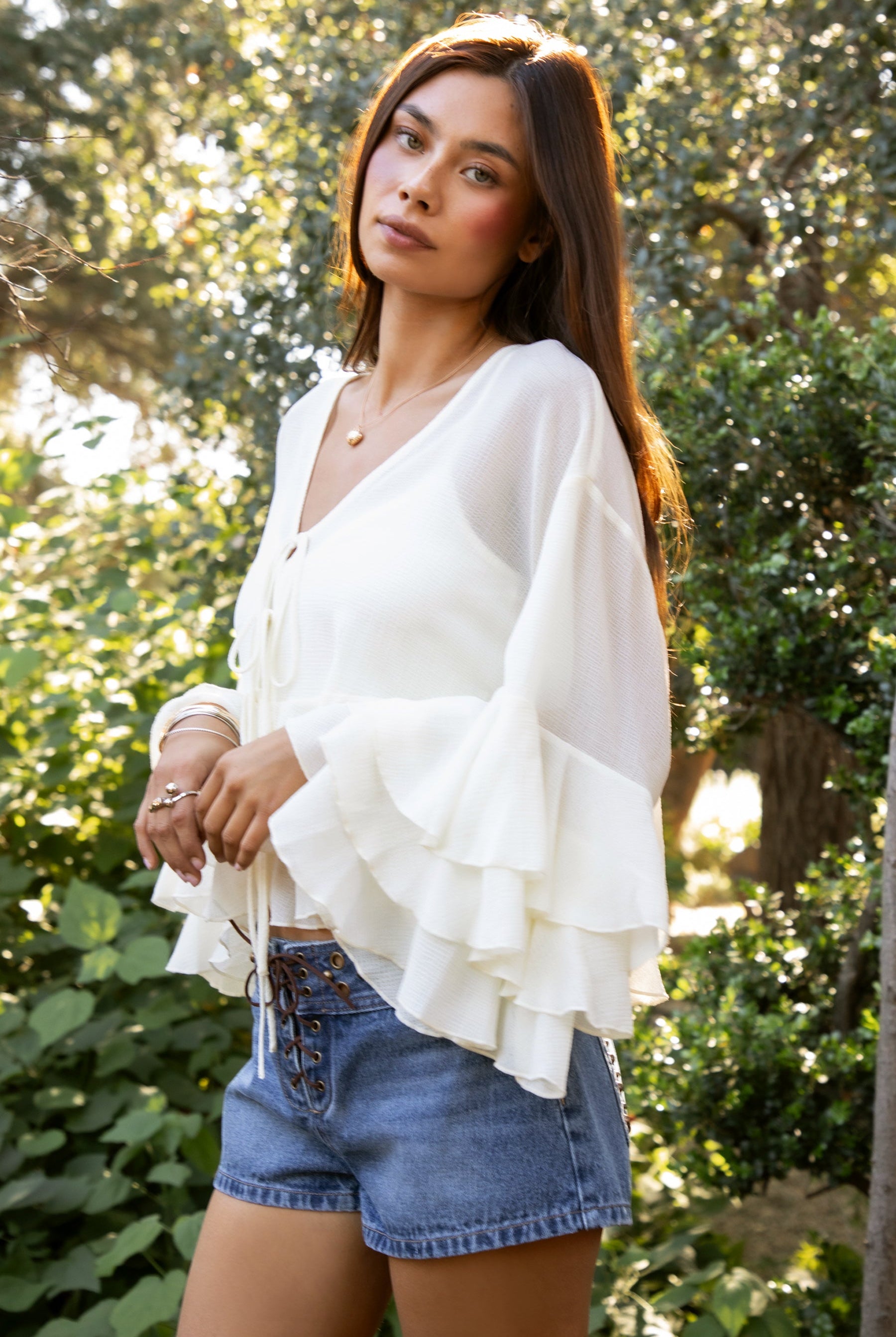 Woman wearing TIC TOC TH31917 BRIEANNE sheer chiffon tiered ruffle blouse featuring a front tie V-neckline, lightweight unlined chiffon body, drop shoulder relaxed fit, and layered ruffle cuffs with a flowy tiered hem, styled for a feminine vacation-ready western look.