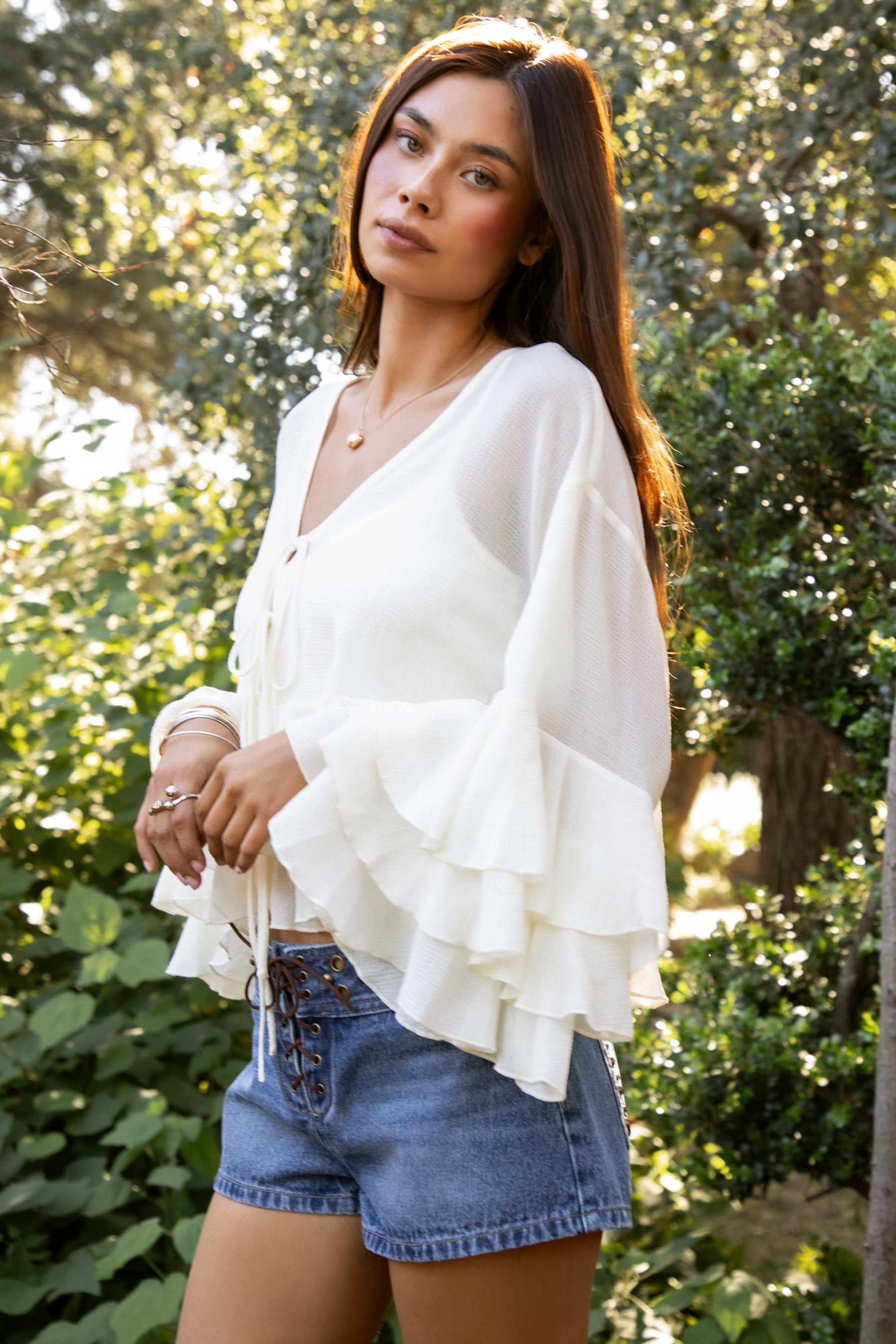 Woman wearing TIC TOC TH31917 BRIEANNE sheer chiffon tiered ruffle blouse featuring a front tie V-neckline, lightweight unlined chiffon body, drop shoulder relaxed fit, and layered ruffle cuffs with a flowy tiered hem, styled for a feminine vacation-ready western look.
