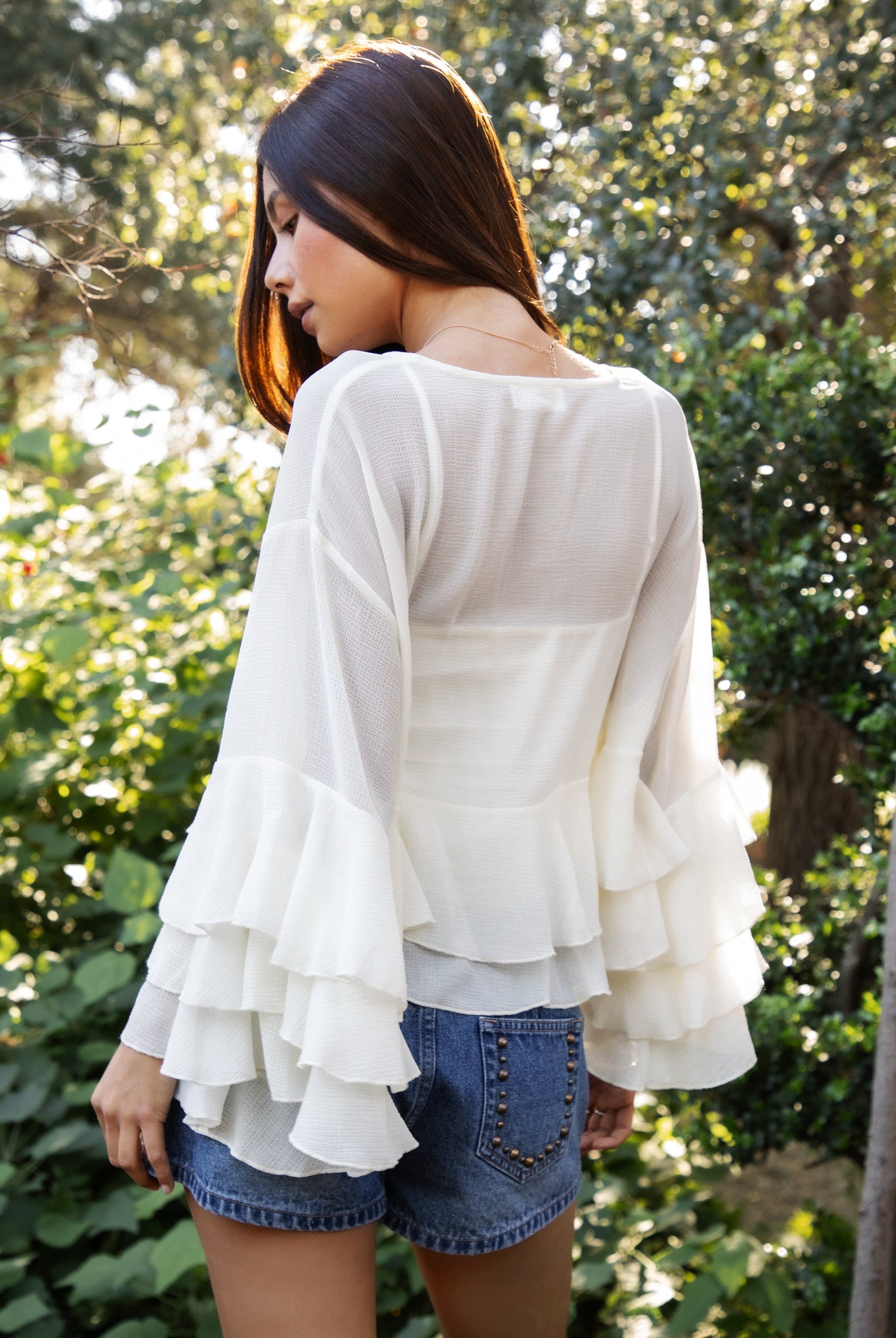 Woman wearing TIC TOC TH31917 BRIEANNE sheer chiffon tiered ruffle blouse featuring a front tie V-neckline, lightweight unlined chiffon body, drop shoulder relaxed fit, and layered ruffle cuffs with a flowy tiered hem, styled for a feminine vacation-ready western look.