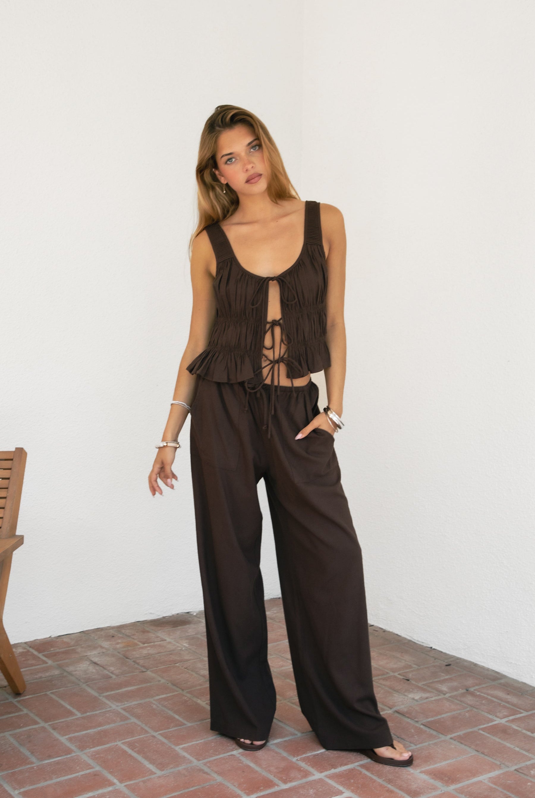Woman wearing SOLSTICE ruched tie front round neck cami top featuring soft ruched texture throughout the body, three self-tie front closures, deep round neckline, wide shoulder straps, and a lightweight viscose linen blend with ruffled hem detail for relaxed resort styling.
