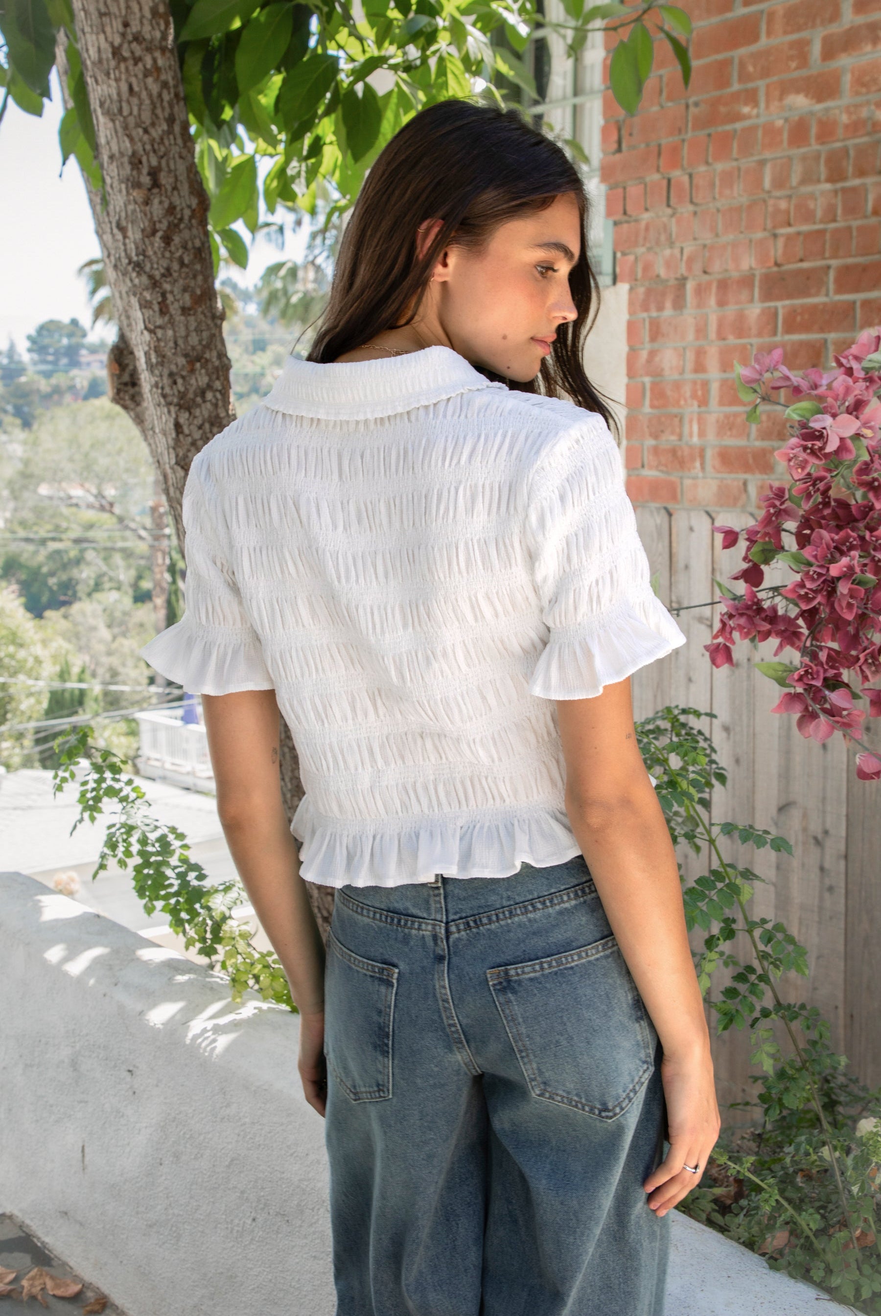 Woman wearing LISETTE smocked button front collared crop top featuring allover smocked textured fabric, soft stretch fit, classic collared neckline, functional button front closure, short puff sleeves with flounce ruffle cuffs, and a cropped silhouette with delicate ruffle hem.