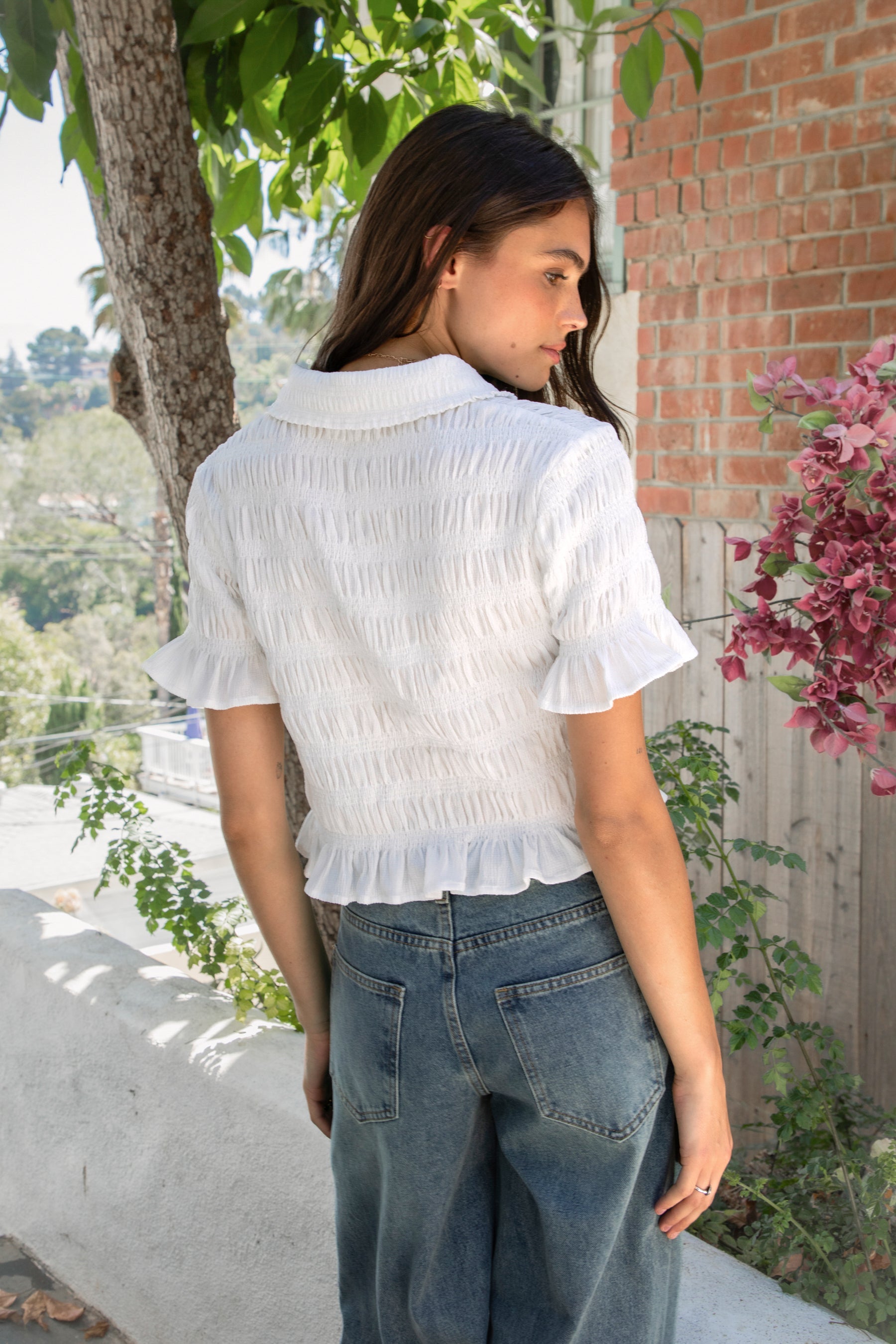 Woman wearing LISETTE smocked button front collared crop top featuring allover smocked textured fabric, soft stretch fit, classic collared neckline, functional button front closure, short puff sleeves with flounce ruffle cuffs, and a cropped silhouette with delicate ruffle hem.