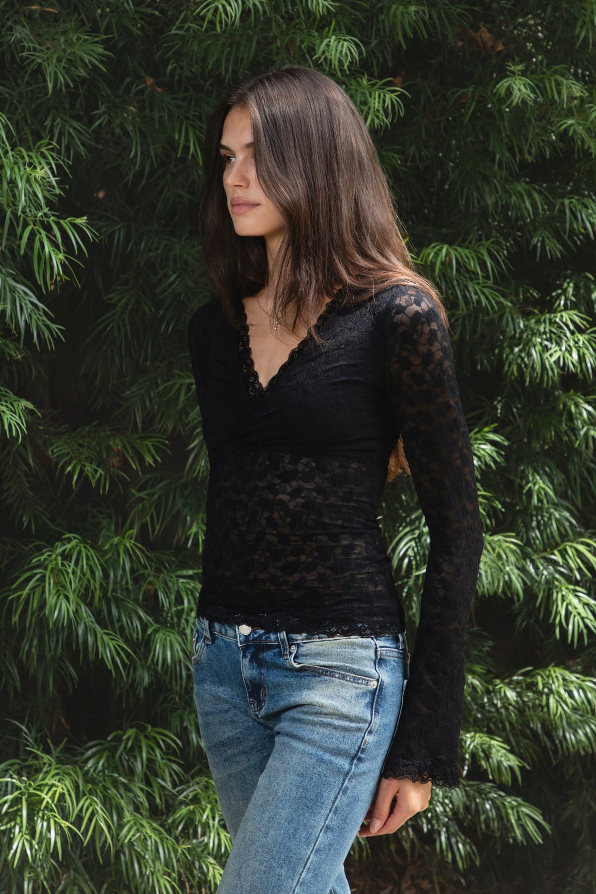 Woman wearing EMMELINE ruched lace long sleeve top featuring sheer floral stretch lace, fitted feminine silhouette, deep V neckline with scalloped lace trim, ruched detailing through the front and sides, soft mesh bust lining, sheer fitted long sleeves, flared lace trimmed cuffs, and scalloped hem finish.