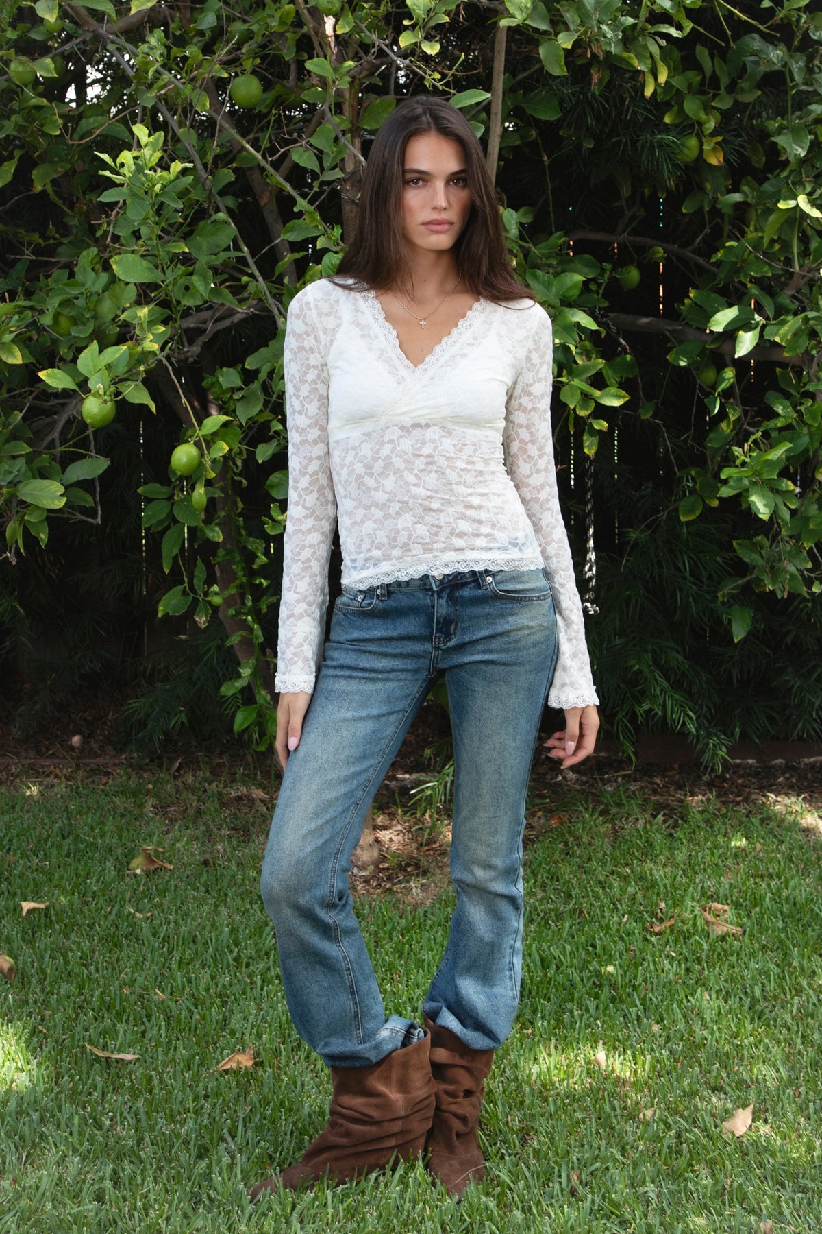 Woman wearing EMMELINE ruched lace long sleeve top featuring sheer floral stretch lace, fitted feminine silhouette, deep V neckline with scalloped lace trim, ruched detailing through the front and sides, soft mesh bust lining, sheer fitted long sleeves, flared lace trimmed cuffs, and scalloped hem finish.