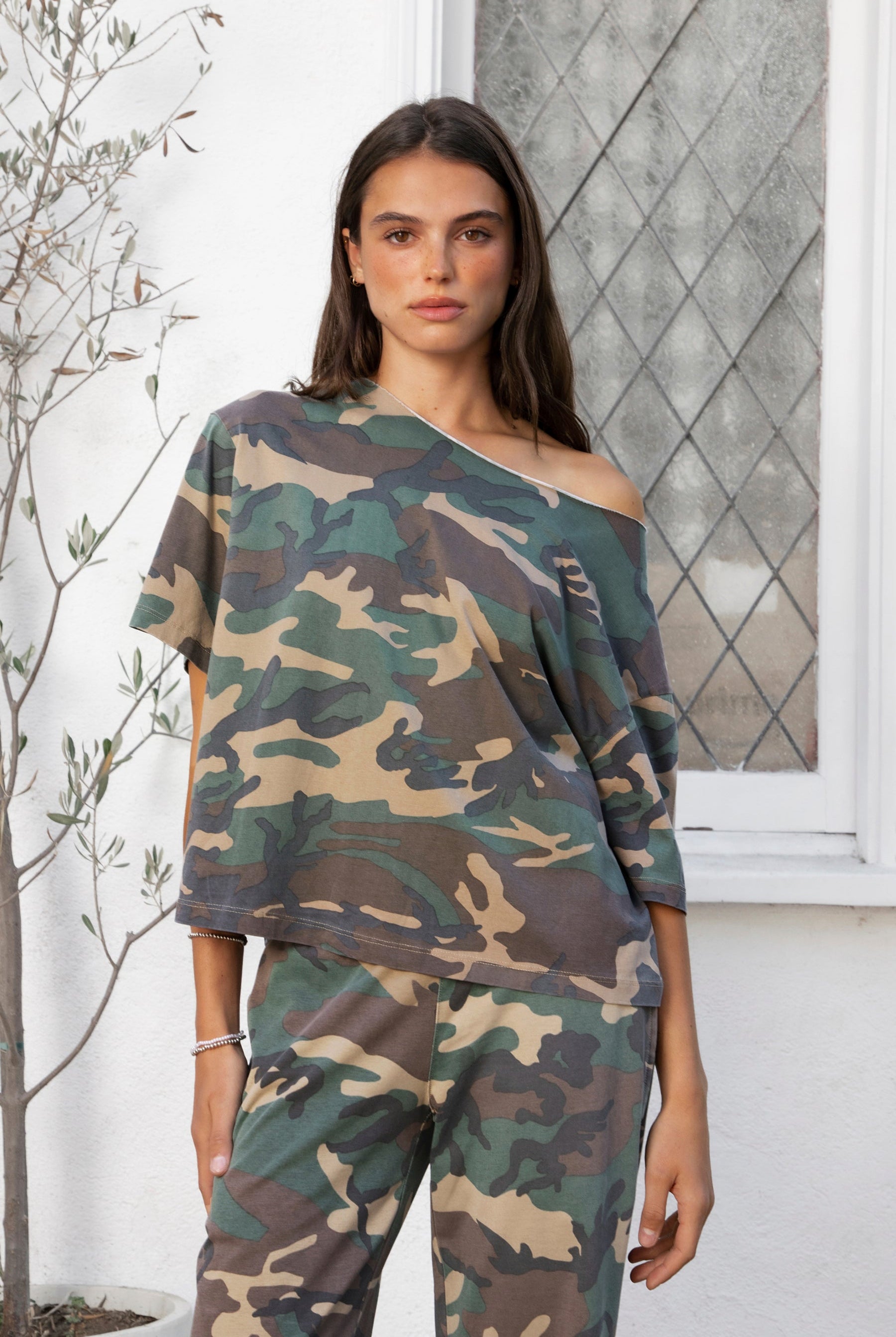 Woman wearing SCOUT military camo short sleeve oversized off shoulder top featuring soft breathable single jersey fabric, allover classic camouflage print, wide rolled raw edge neckline, relaxed oversized silhouette, dropped shoulders, elbow length sleeves, and casual boyfriend inspired draped fit.