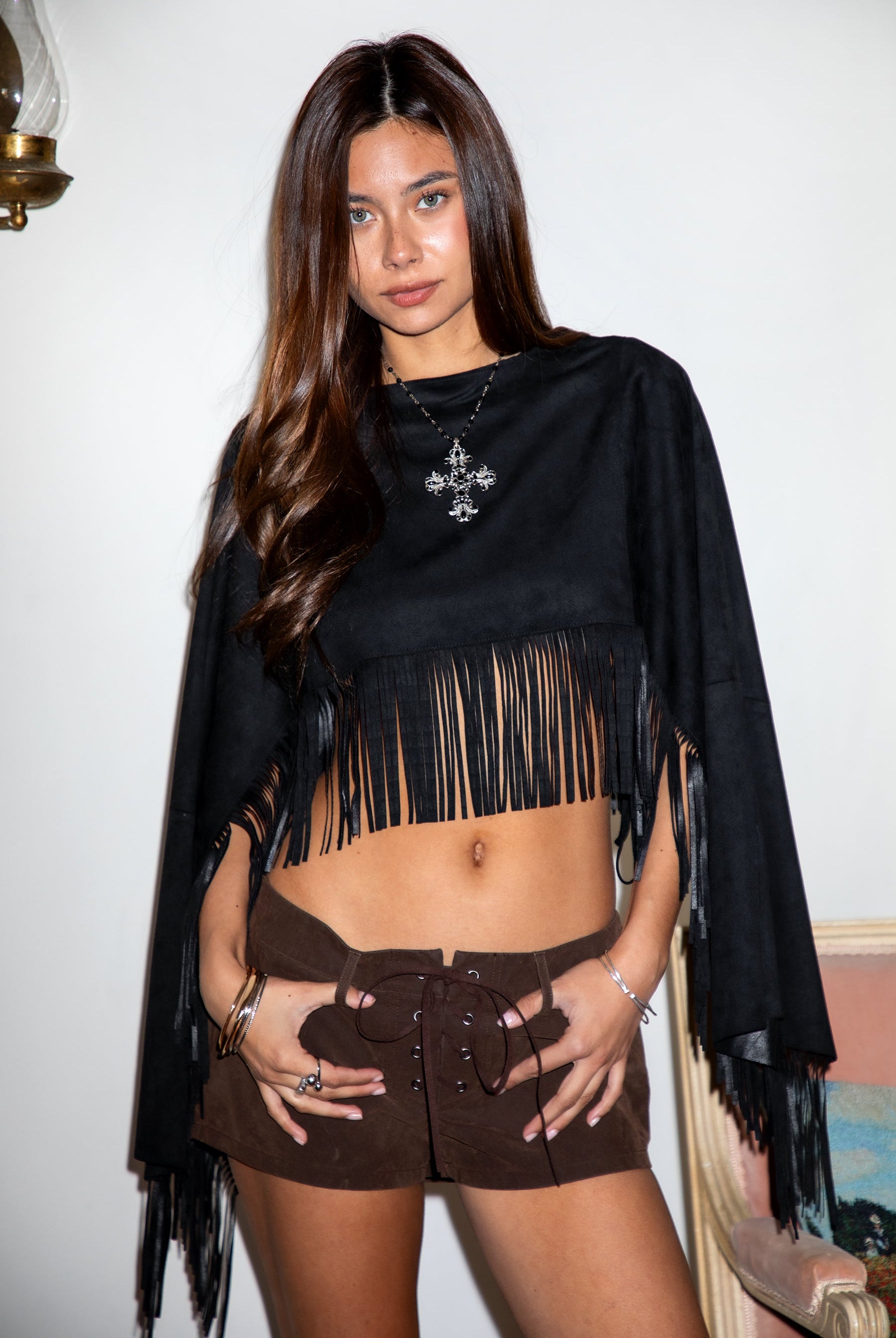 Woman wearing TIC TOC TL31825 STALLION faux suede fringe poncho featuring a cropped cape silhouette, drop shoulder relaxed fit, long fringe tassel detailing along the hem and sleeves, and a soft western-inspired faux suede texture, styled over a fitted top with denim bottoms for a rodeo festival look.