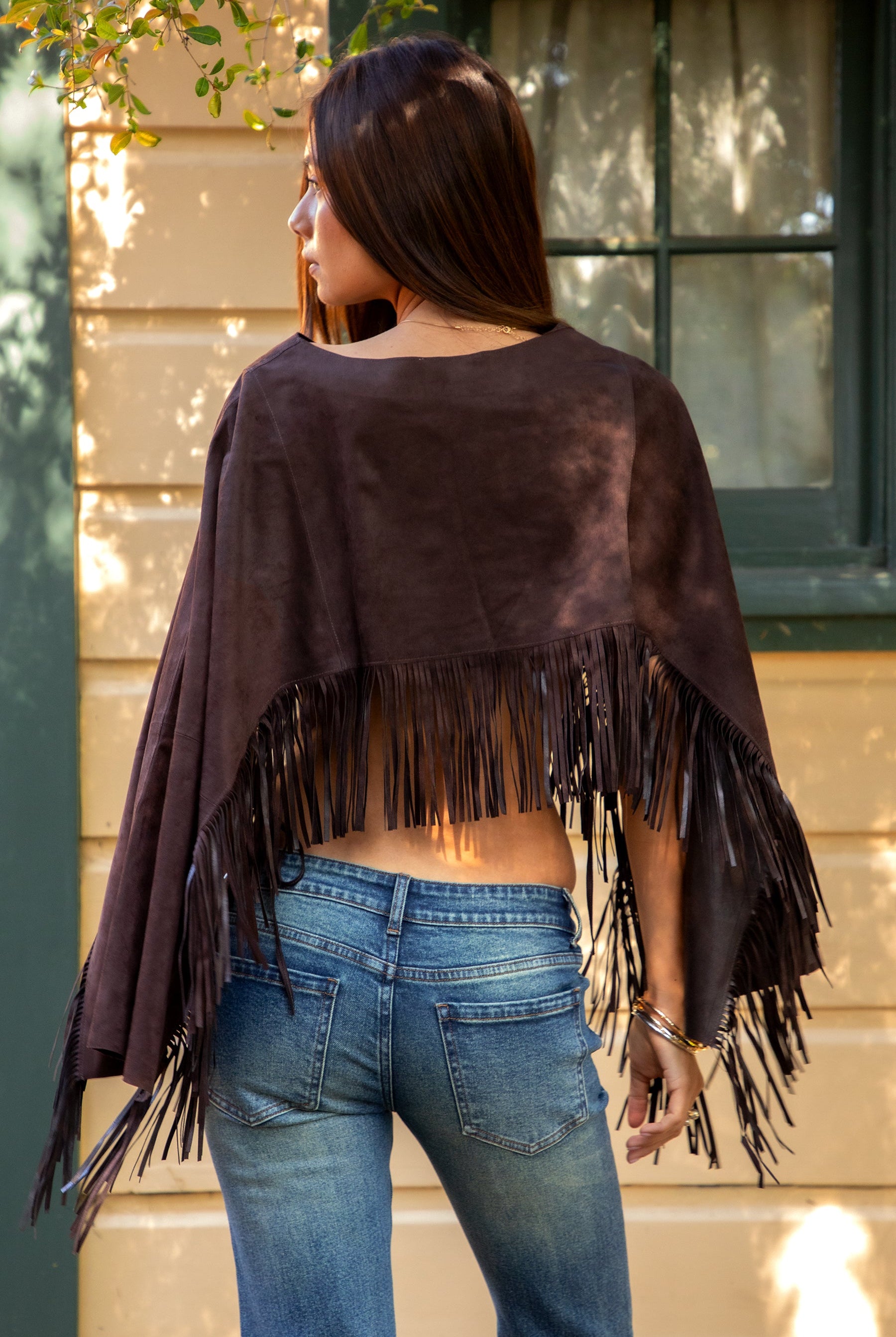 Woman wearing TIC TOC TL31825 STALLION faux suede fringe poncho featuring a cropped cape silhouette, drop shoulder relaxed fit, long fringe tassel detailing along the hem and sleeves, and a soft western-inspired faux suede texture, styled over a fitted top with denim bottoms for a rodeo festival look.