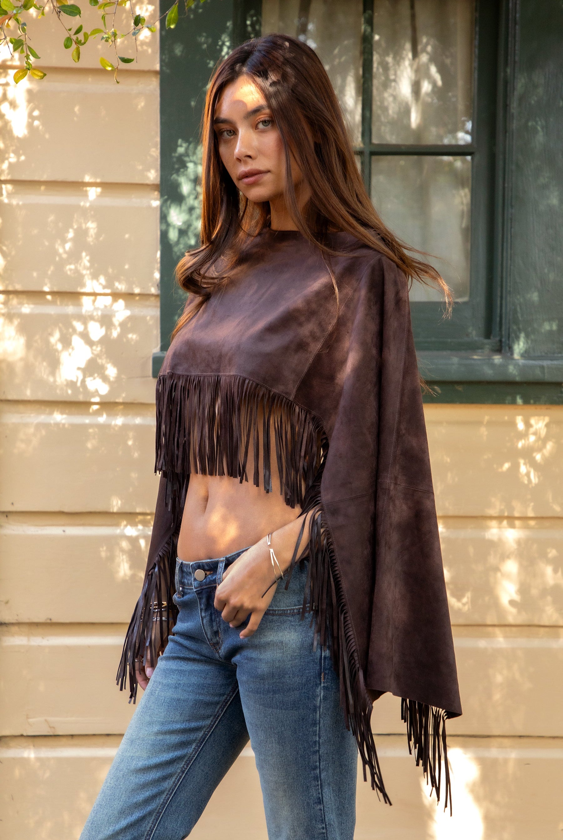 Woman wearing TIC TOC TL31825 STALLION faux suede fringe poncho featuring a cropped cape silhouette, drop shoulder relaxed fit, long fringe tassel detailing along the hem and sleeves, and a soft western-inspired faux suede texture, styled over a fitted top with denim bottoms for a rodeo festival look.