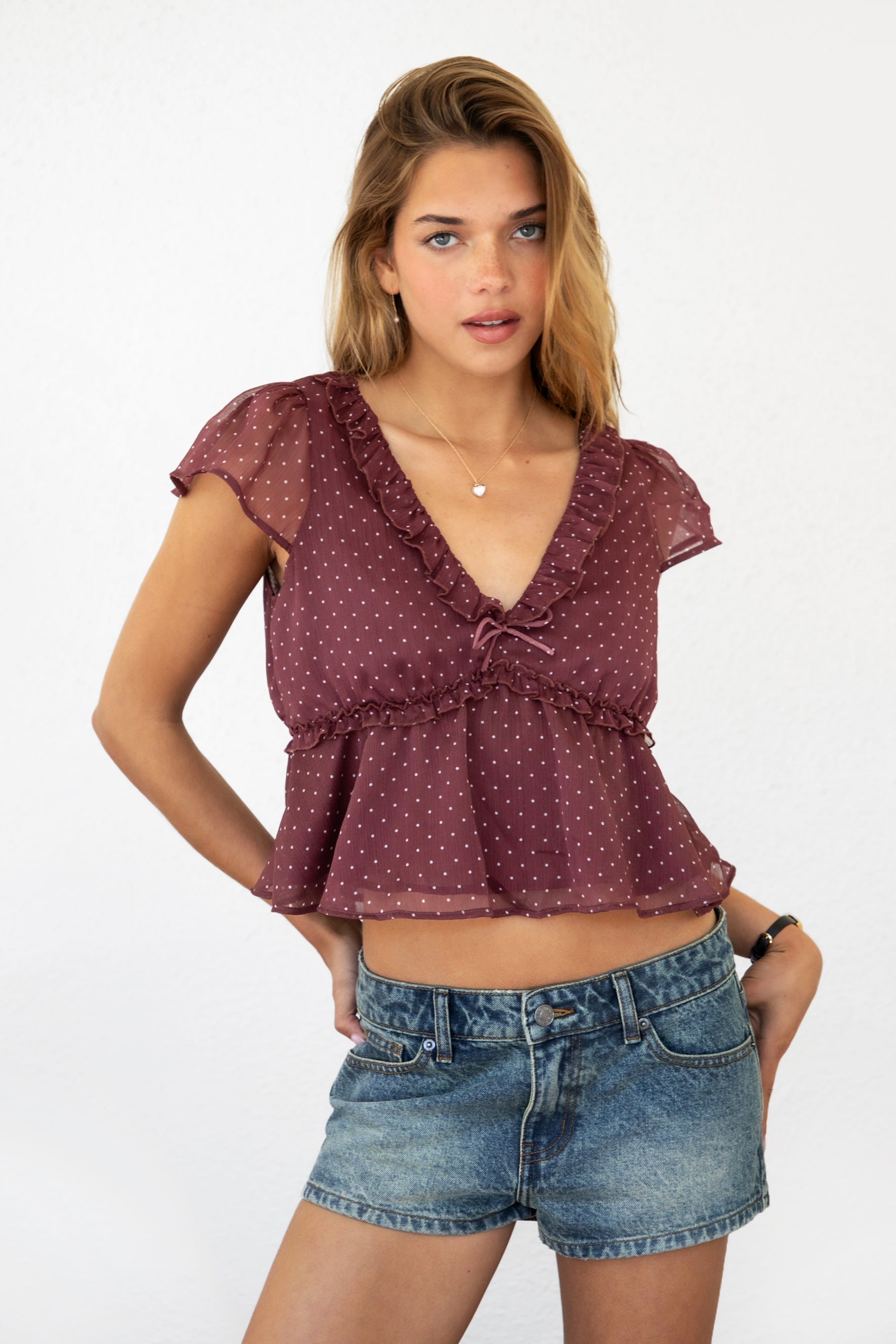 Woman wearing WYNONA polka dot chiffon crop top featuring lightweight printed chiffon, V-neckline with ruffle trim, flutter sleeves, elastic underbust band, front bow detail, and a cropped peplum silhouette with full lining for comfort.