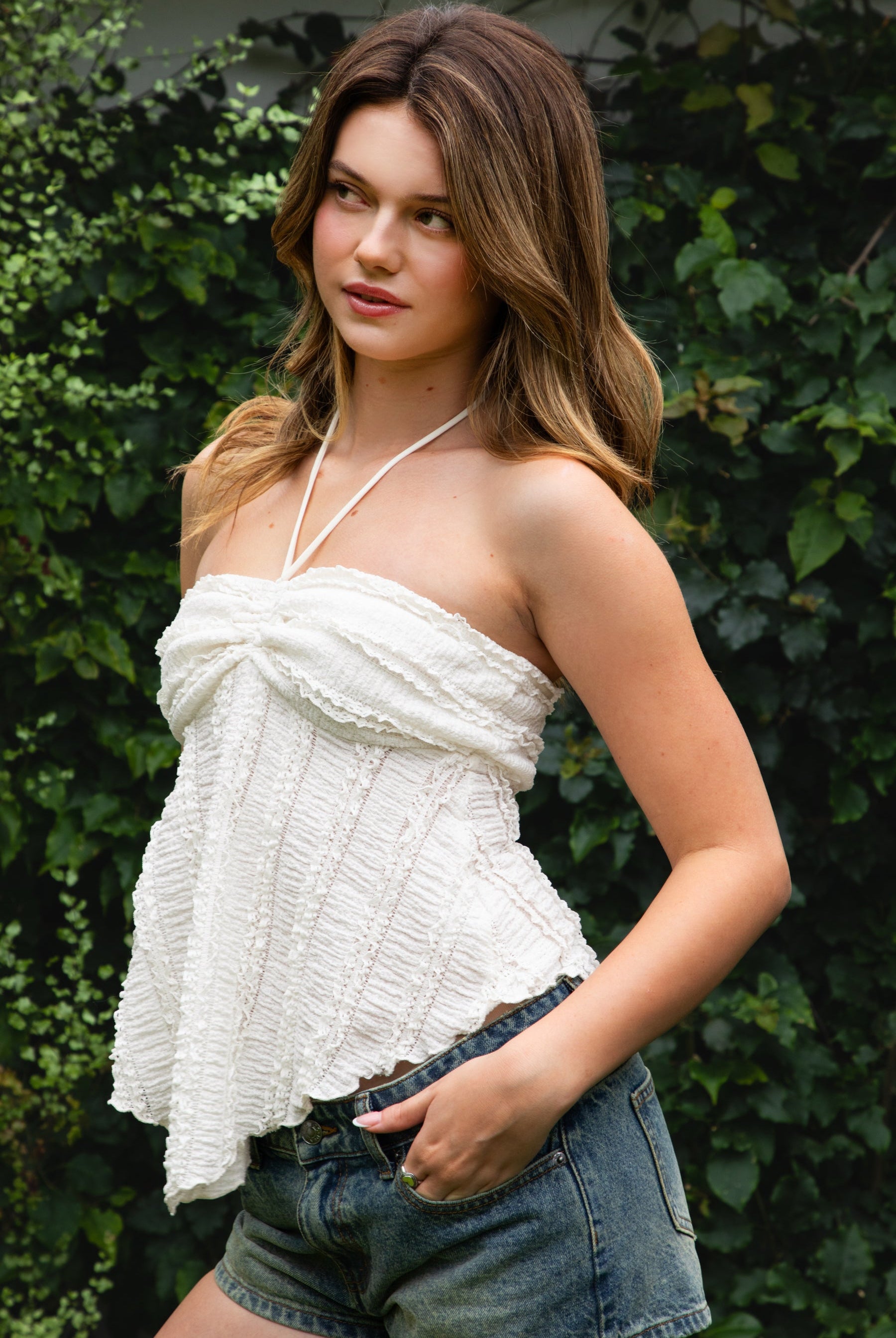 Woman wearing ODETTE lace handkerchief halter top featuring soft textured lace fabric, twisted bandeau bodice, adjustable halter neck tie, romantic handkerchief hemline, delicate lace trim, ruched texture, and lightweight lining for summer vacation and festival styling.