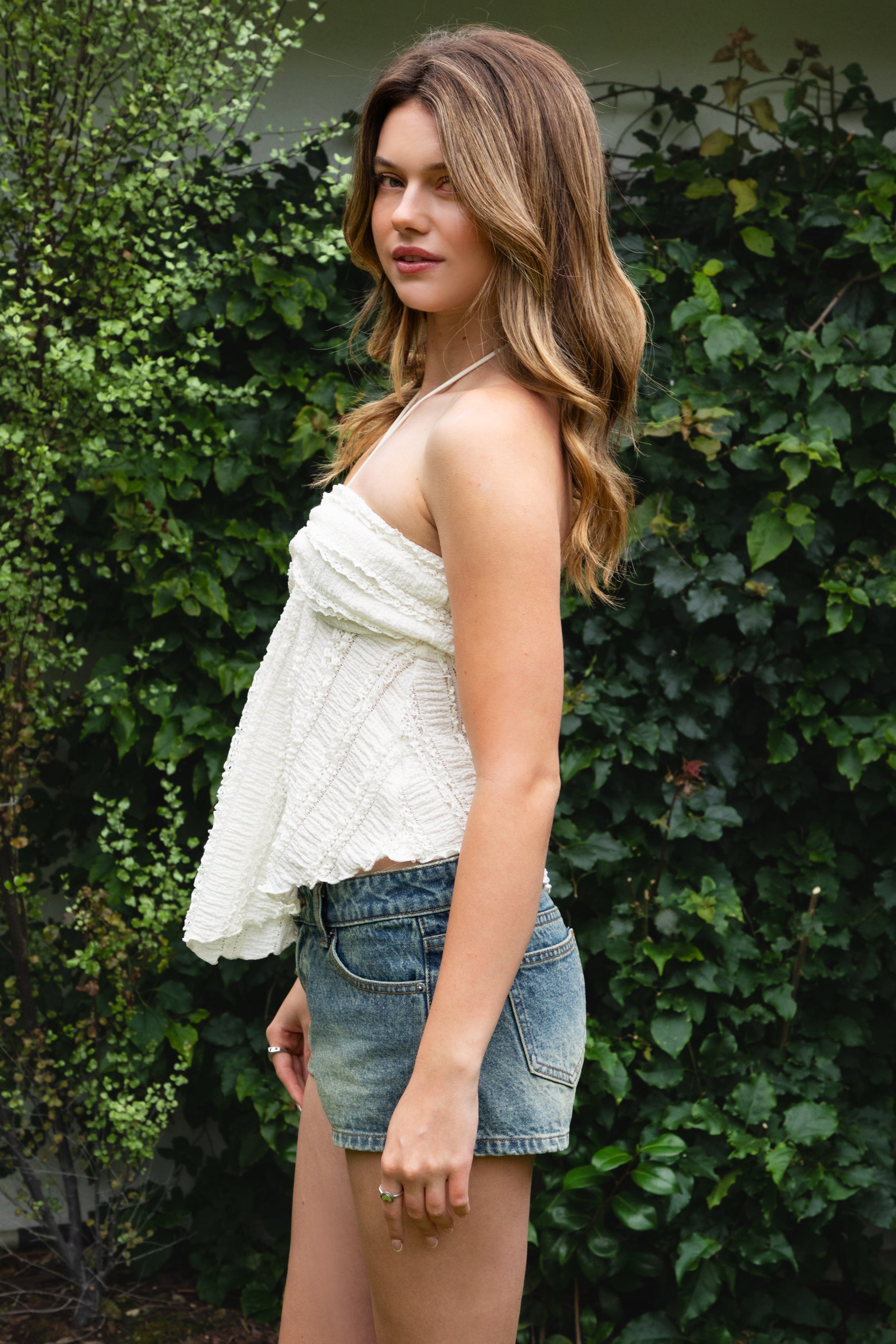 Woman wearing ODETTE lace handkerchief halter top featuring soft textured lace fabric, twisted bandeau bodice, adjustable halter neck tie, romantic handkerchief hemline, delicate lace trim, ruched texture, and lightweight lining for summer vacation and festival styling.