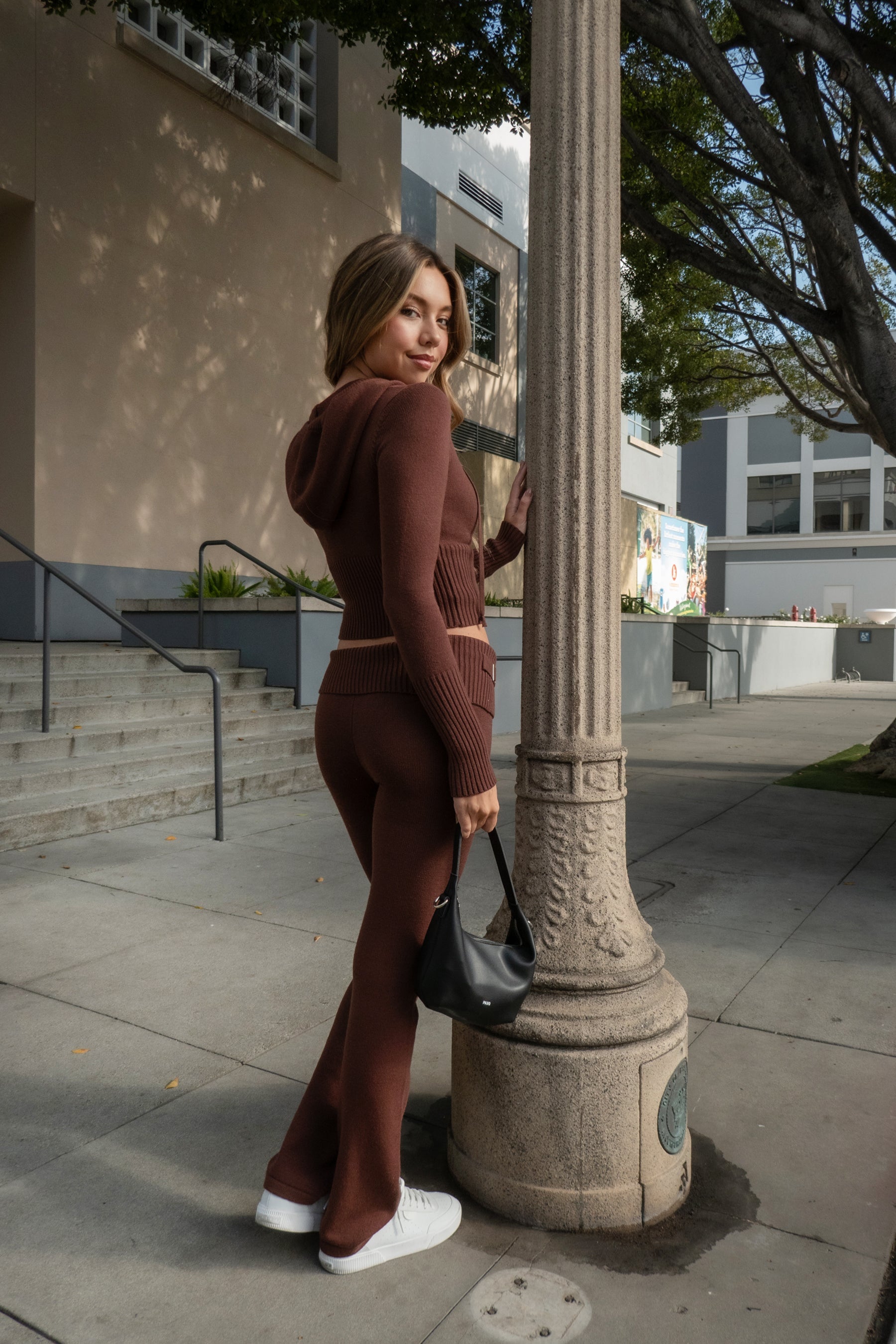 Woman wearing TIC TOC TPC29297-COZEE brown sweater knit two-piece set featuring a cropped zip-up hoodie with ribbed details and matching fold-over flare pants, styled outdoors with sneakers and a small black shoulder bag.