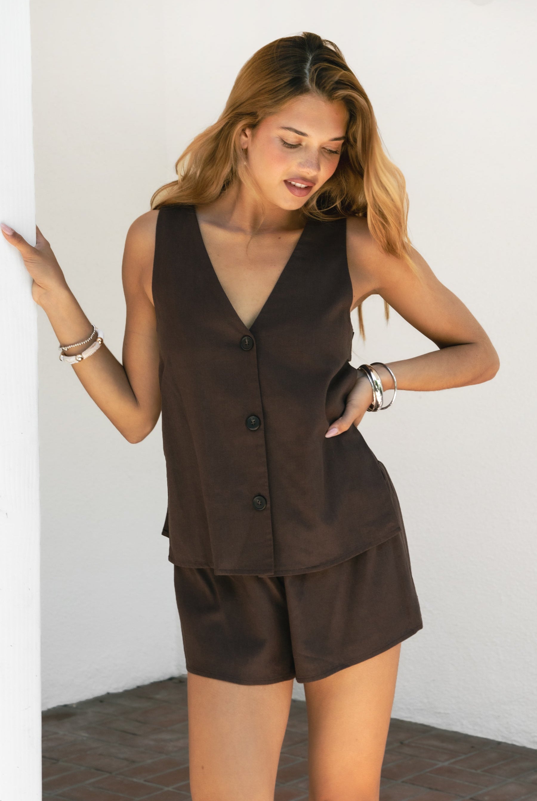 Woman wearing ELENI viscose linen sleeveless top and shorts set featuring a relaxed V-neck vest with three-button front closure, split side detail with dual buttons, and easy pull-on shorts with elastic waistband in a lightweight breathable linen blend.