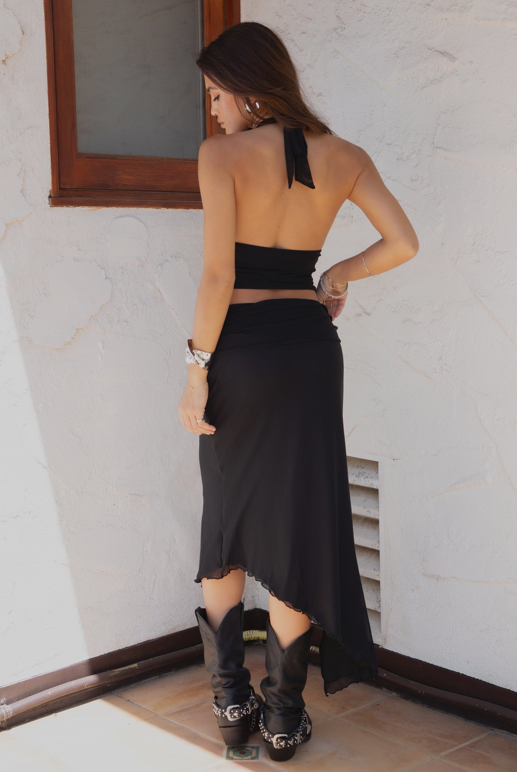 Woman wearing TIC TOC ALMEDA Mesh Print Halter and Maxi Skirt Set in black with studded detail, scarf hem top, and high slit skirt paired with black cowboy boots against a white wall.