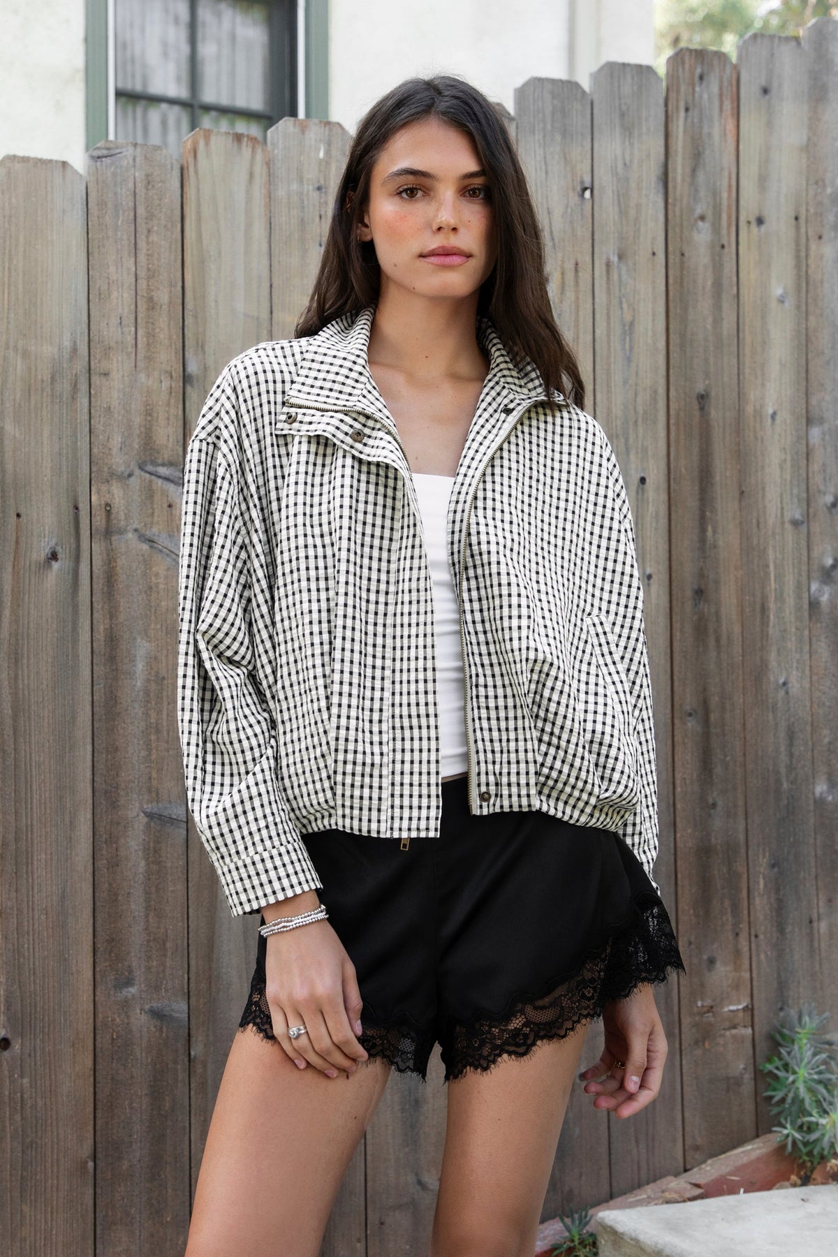 Woman wearing MARGOT plaid gingham zip up bomber jacket featuring lightweight crinkled cotton blend fabric, classic gingham plaid pattern, high neck collar, front button and zip closure, semi cropped length, sleeve snap buttons, relaxed drop shoulder silhouette, side pockets, and elastic hem.
