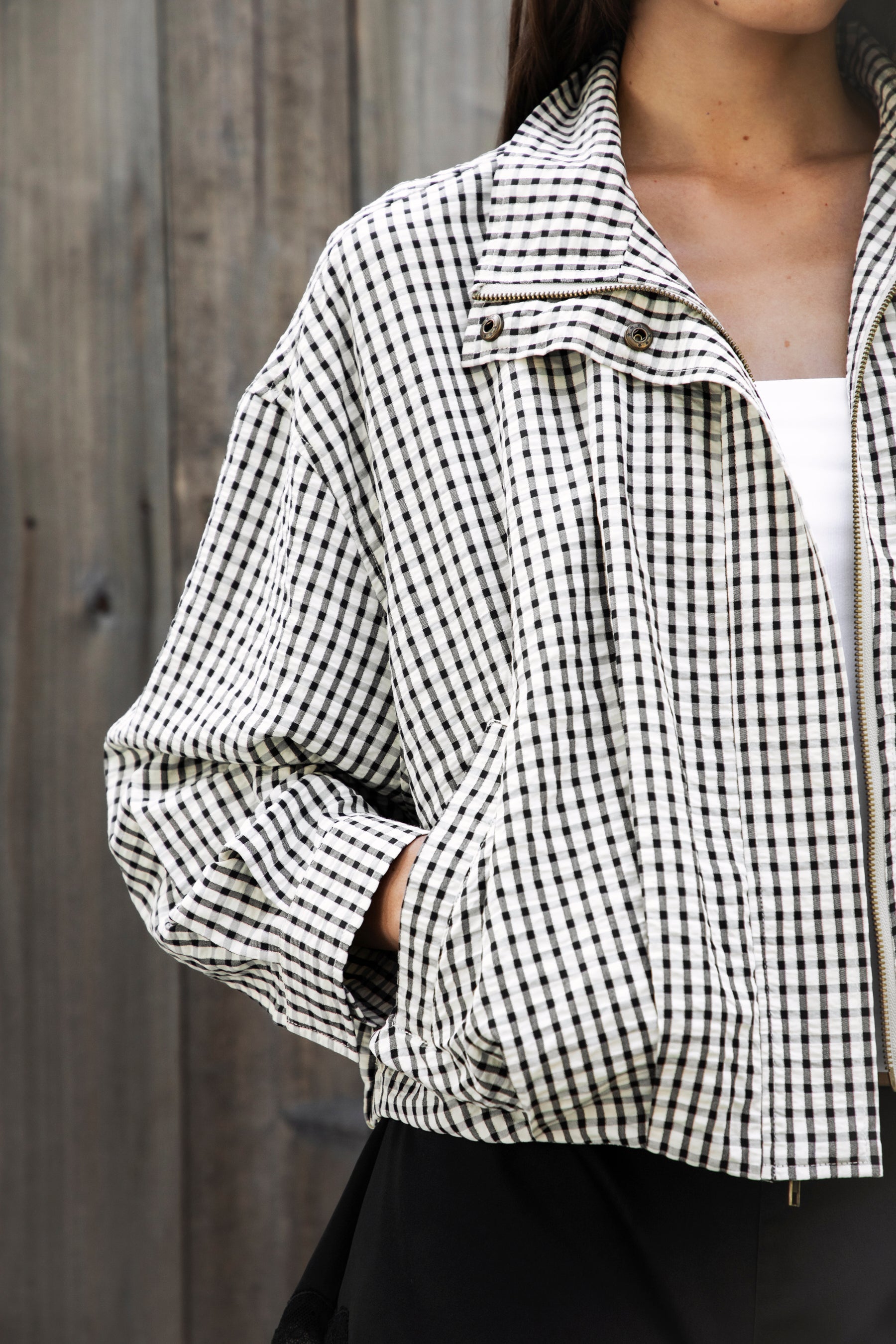 Woman wearing MARGOT plaid gingham zip up bomber jacket featuring lightweight crinkled cotton blend fabric, classic gingham plaid pattern, high neck collar, front button and zip closure, semi cropped length, sleeve snap buttons, relaxed drop shoulder silhouette, side pockets, and elastic hem.