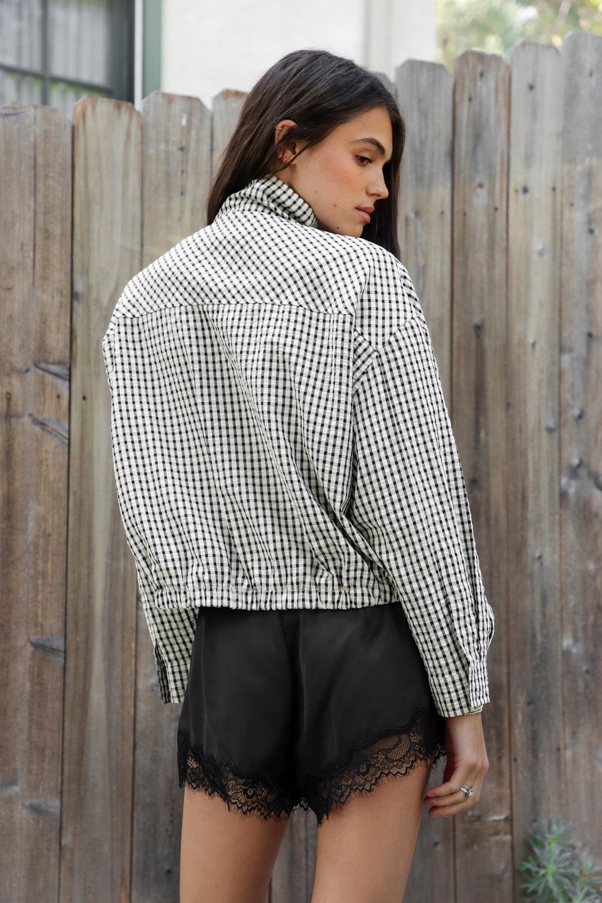 Woman wearing MARGOT plaid gingham zip up bomber jacket featuring lightweight crinkled cotton blend fabric, classic gingham plaid pattern, high neck collar, front button and zip closure, semi cropped length, sleeve snap buttons, relaxed drop shoulder silhouette, side pockets, and elastic hem.