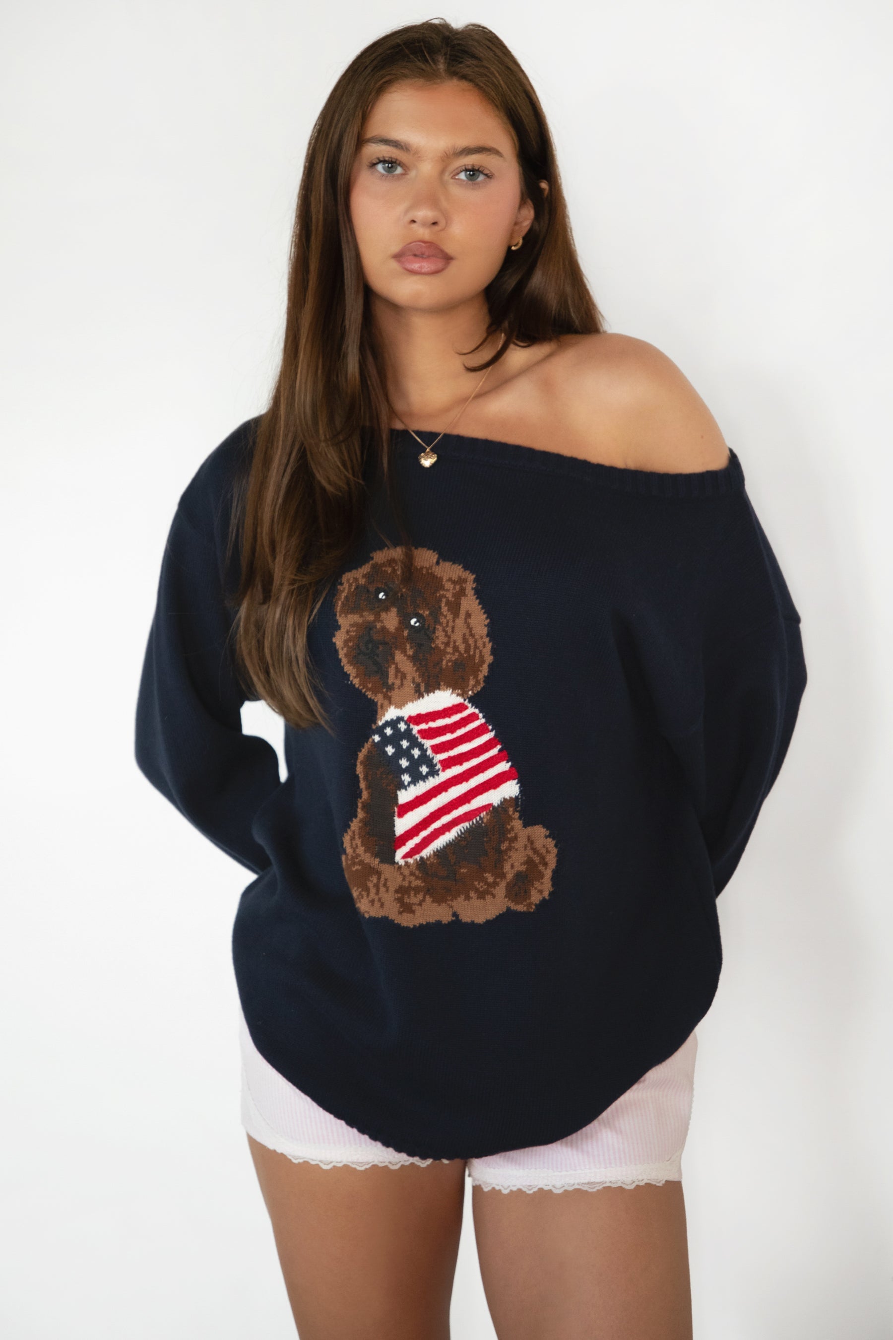 Woman wearing TIC TOC TV31893-MALTIE MALTIE knit sweater featuring a wide boat off-the-shoulder neckline, oversized relaxed fit, dropped shoulders, and long sleeves with ribbed cuffs, showcasing a poodle intarsia front graphic with American flag detailing, styled casually with denim for a cozy Americana-inspired weekend look.