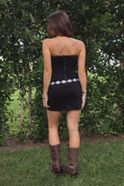 Woman wearing a black dress with silver embellishments and brown boots standing in a grassy area with trees.