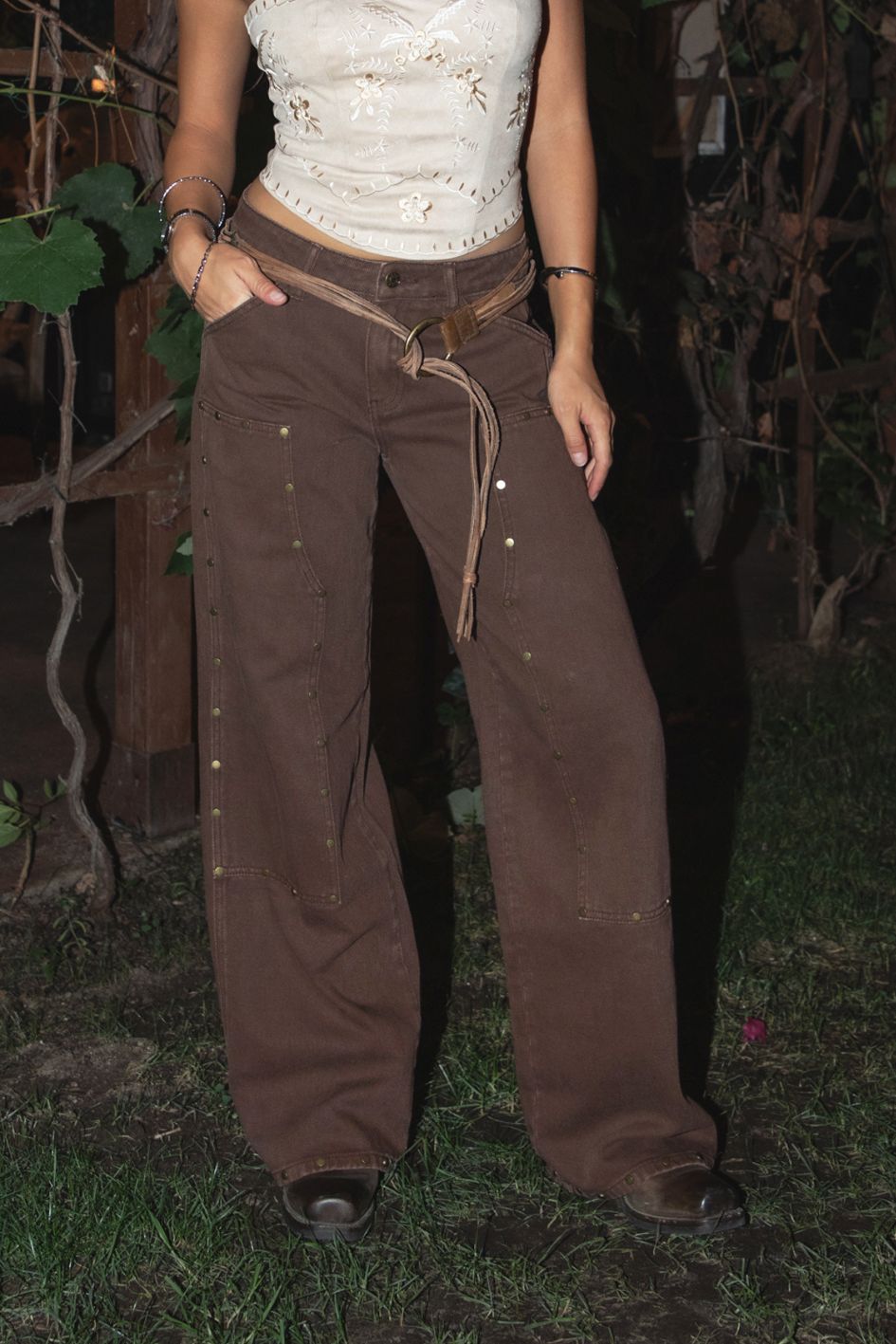 Woman wearing the TIC TOC PF31461 IZLA brown denim twill mid rise studded carpenter pants featuring double-knee seams, wide-leg fit, and vintage faded wash paired with an embroidered cream strapless top in an outdoor setting.