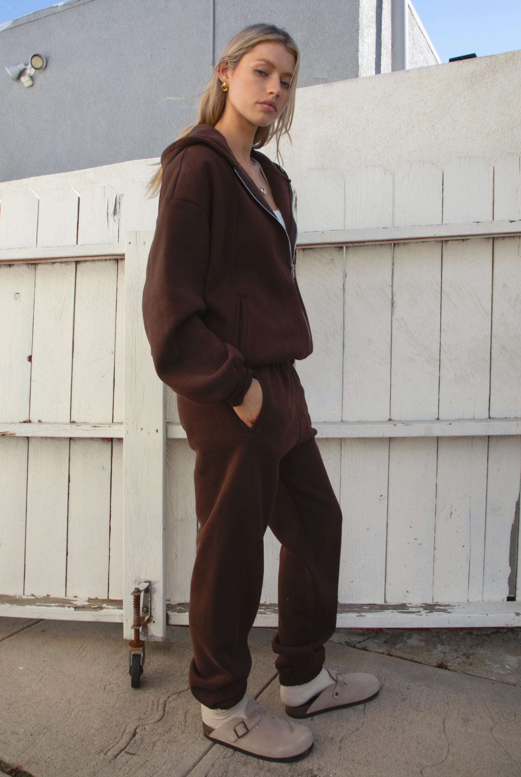 Person wearing a brown tracksuit and white crop top in front of a white wooden door.