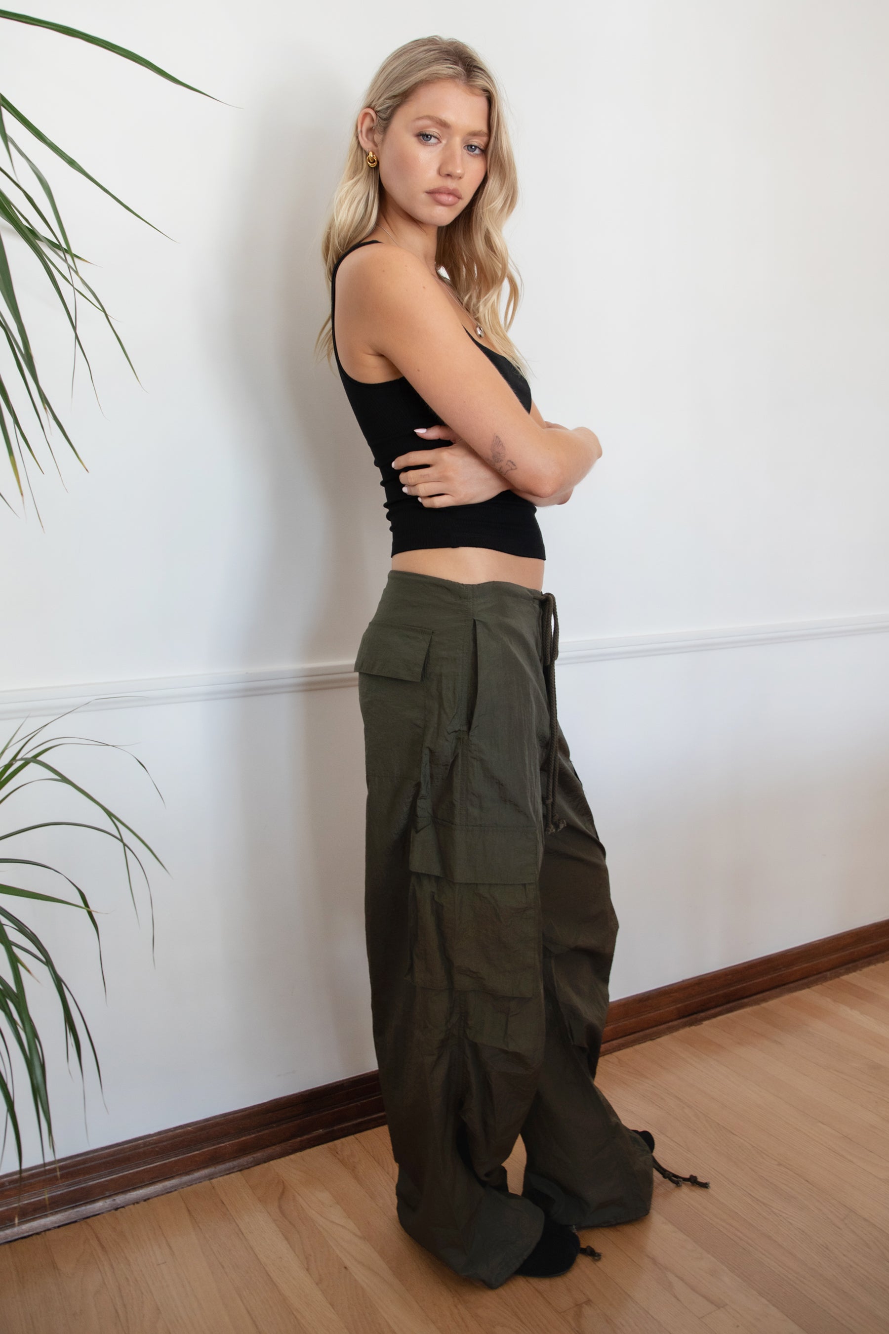 Woman wearing a black tank top and TIC TOC PH29131 TOMY olive crinkled nylon parachute cargo pants with adjustable drawstring waistband and wide-leg silhouette against a white wall.