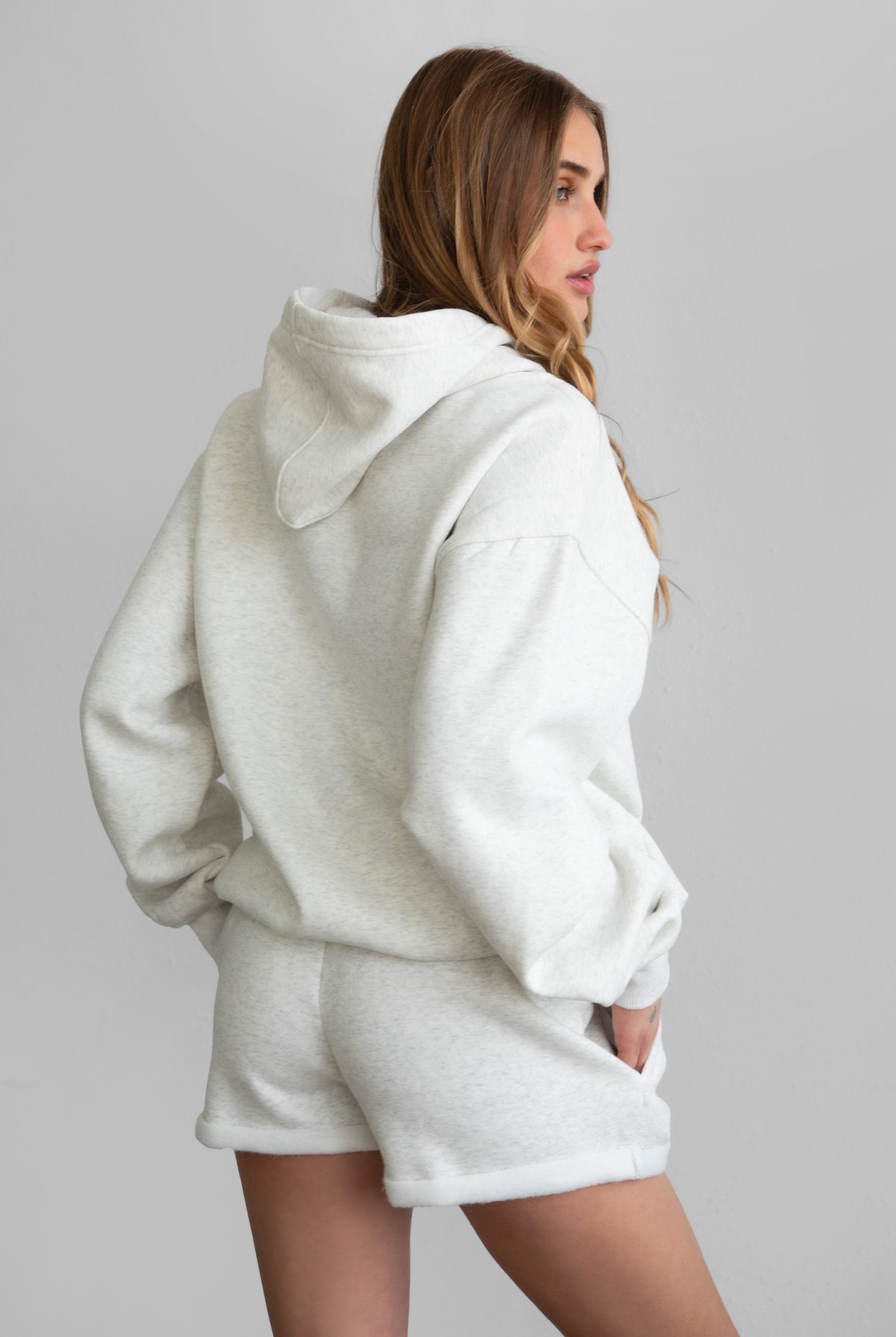 Woman wearing the TIC TOC PJ31004 CARLY heather white cloud fleece low rise shorts featuring an elastic waistband with drawcord, deep side pockets, and raw cut hemline paired with a matching top