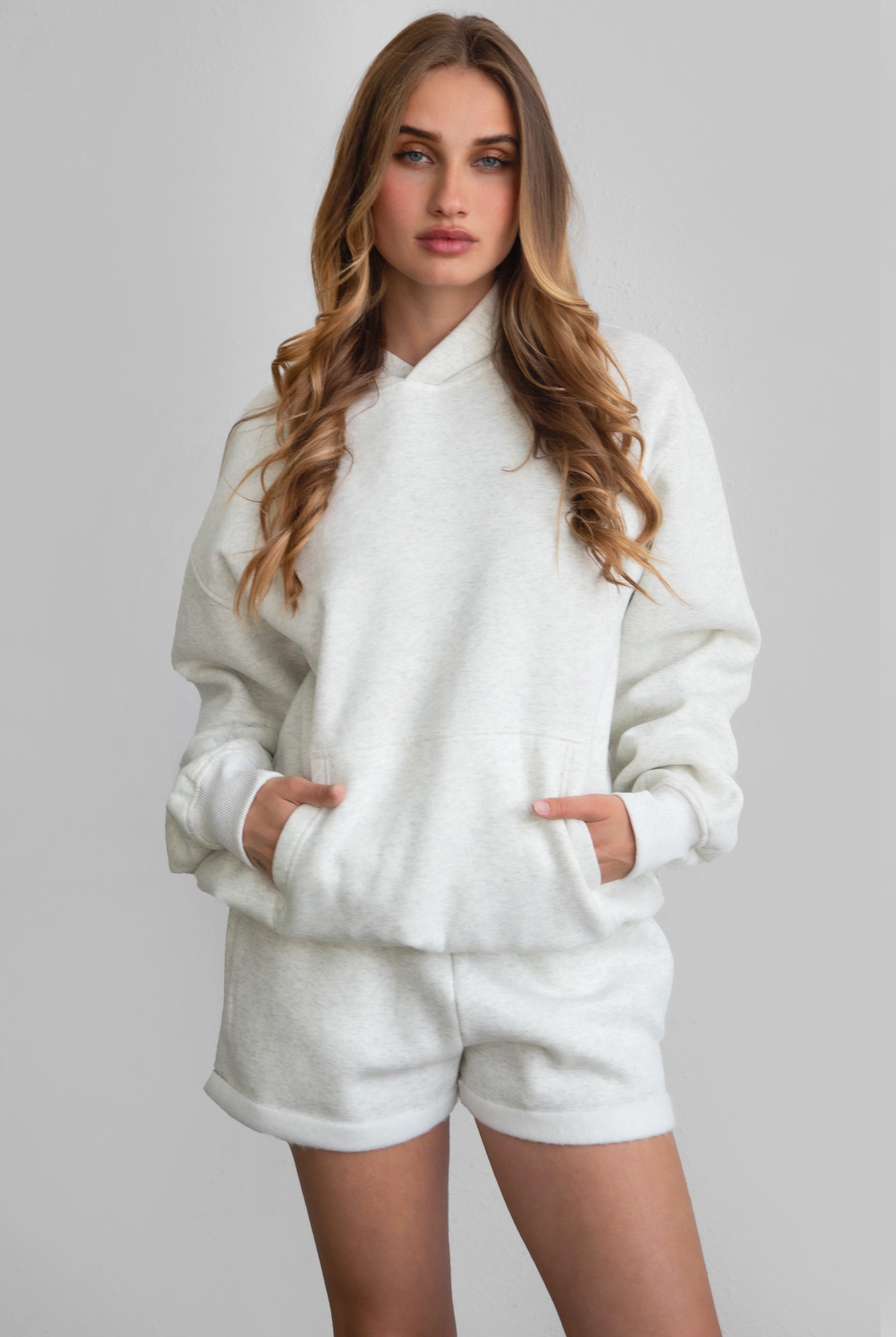 Woman wearing the TIC TOC PJ31004 CARLY heather white cloud fleece low rise shorts featuring an elastic waistband with drawcord, deep side pockets, and raw cut hemline paired with a matching top