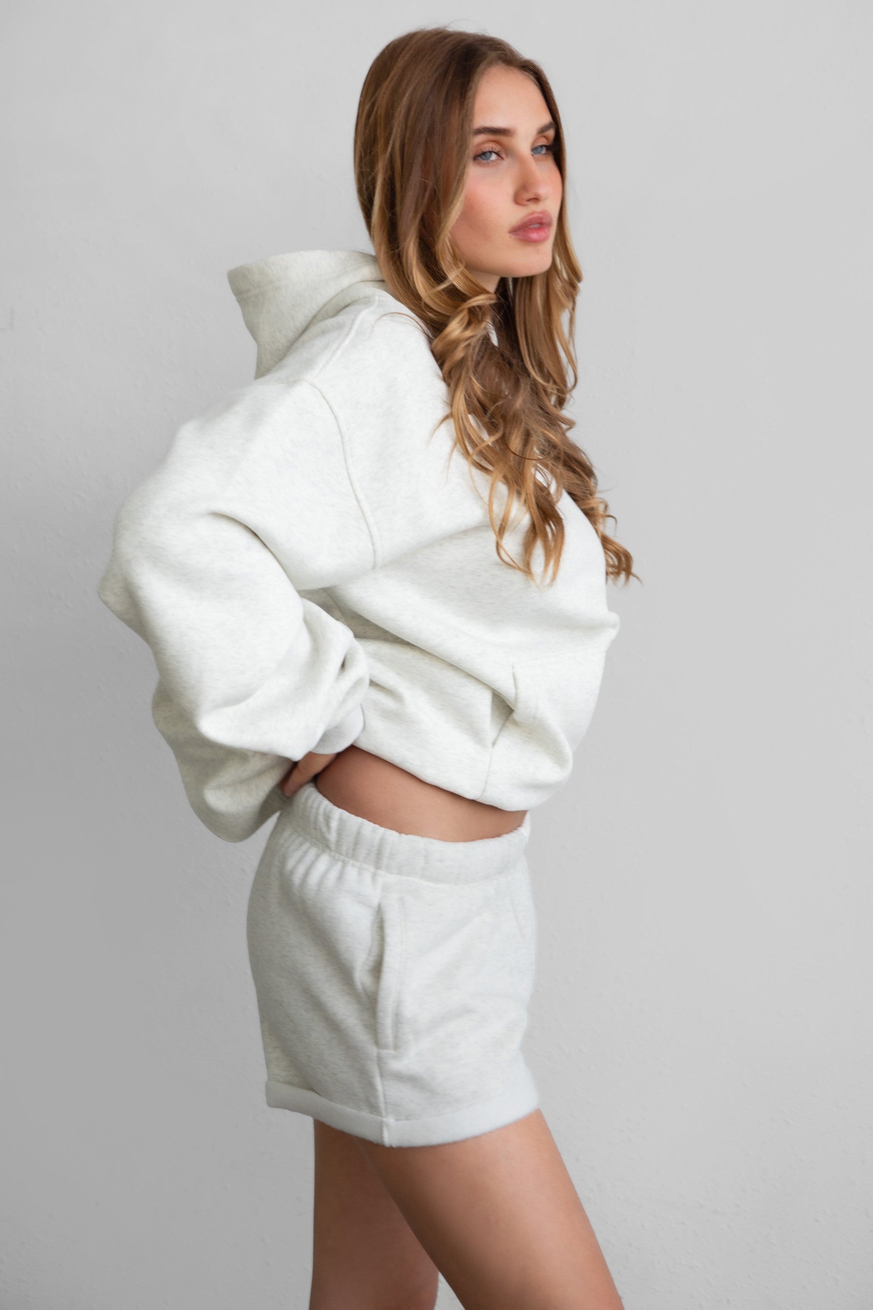 Woman wearing the TIC TOC PJ31004 CARLY heather white cloud fleece low rise shorts featuring an elastic waistband with drawcord, deep side pockets, and raw cut hemline paired with a matching top