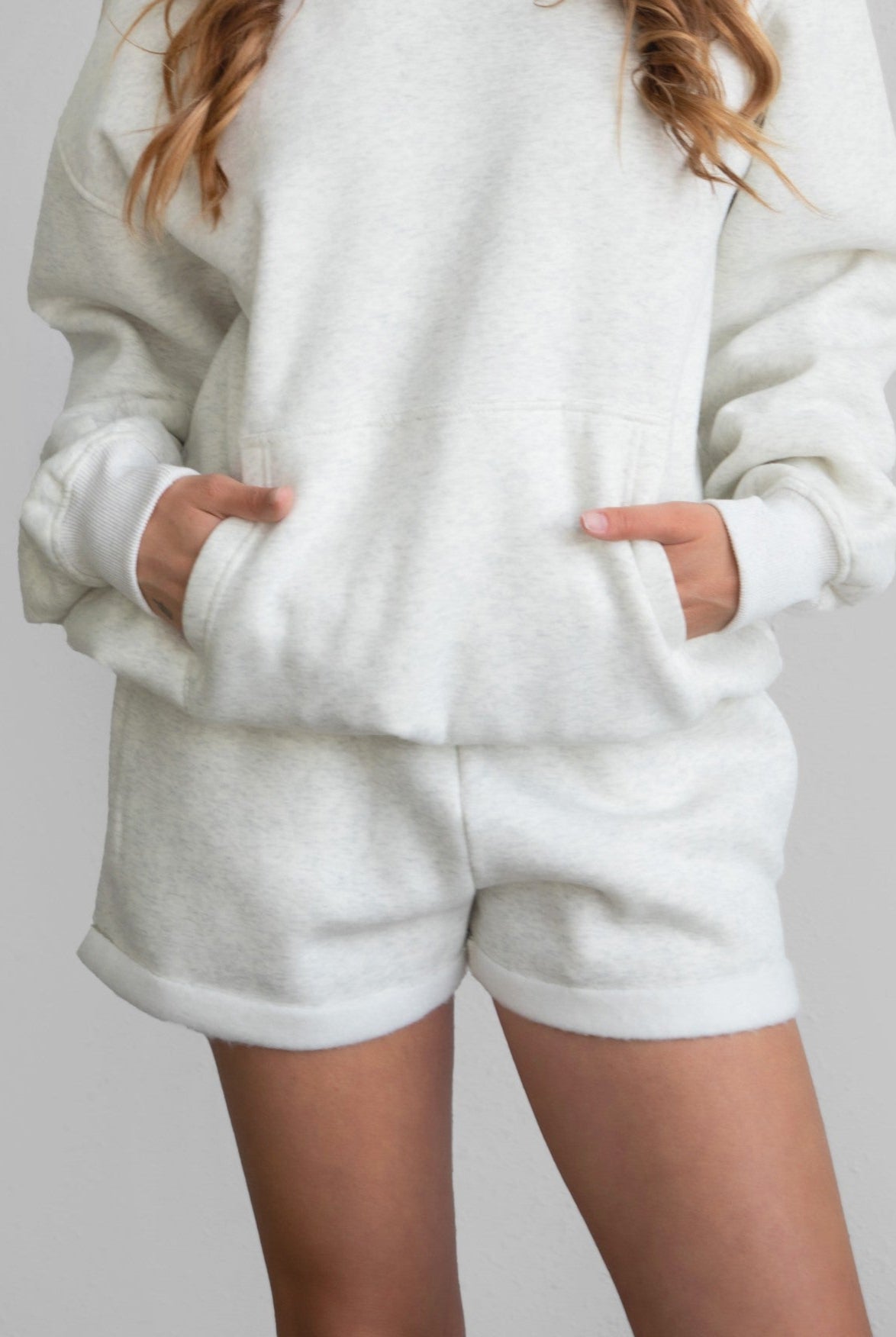 Woman wearing the TIC TOC PJ31004 CARLY heather white cloud fleece low rise shorts featuring an elastic waistband with drawcord, deep side pockets, and raw cut hemline paired with a matching top