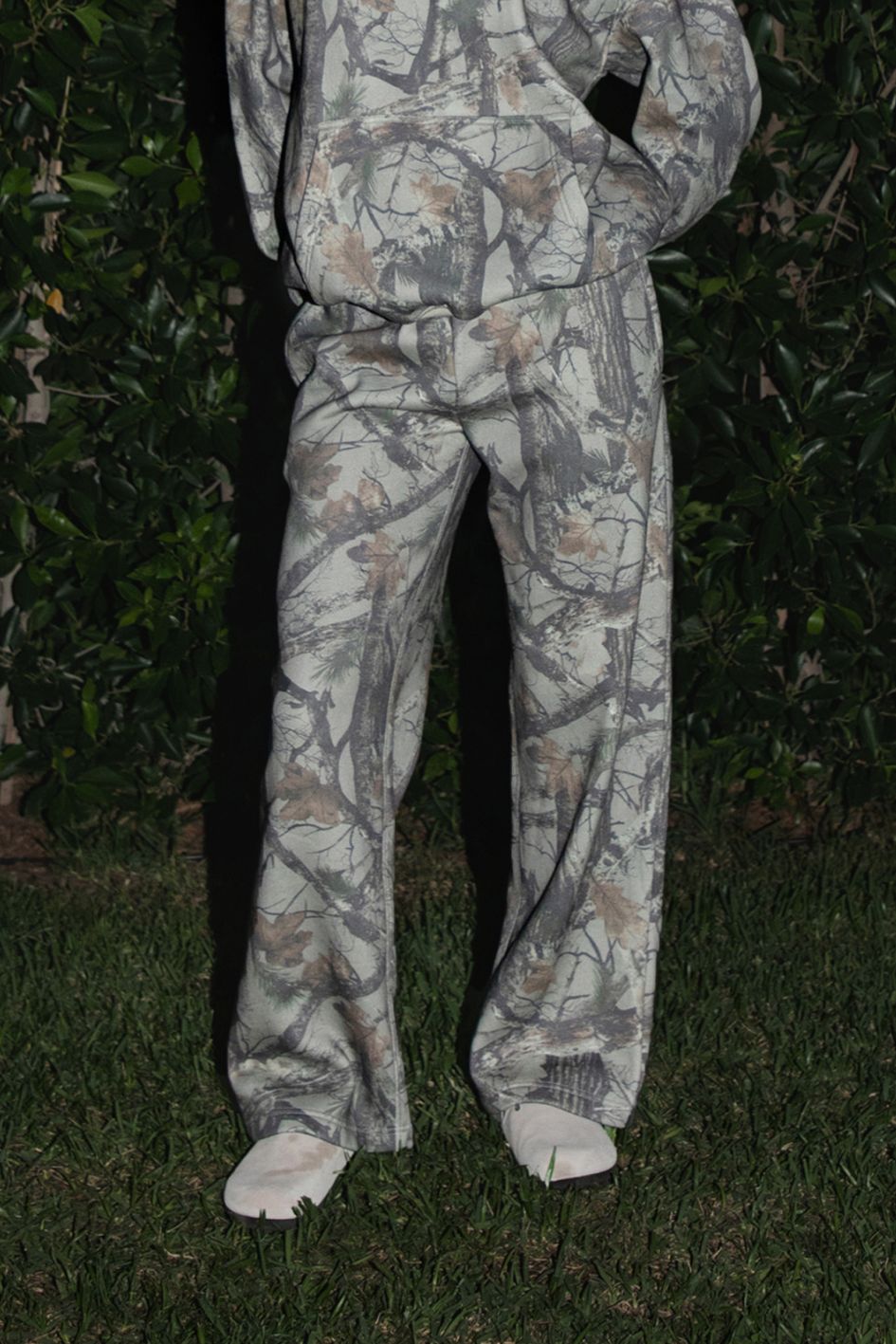 Woman wearing TIC TOC PJ31369 ELK woodland camo fleece wide leg sweatpants with elastic waistband, drawcord, and side pockets with matching top standing in grass with a bush in the background
