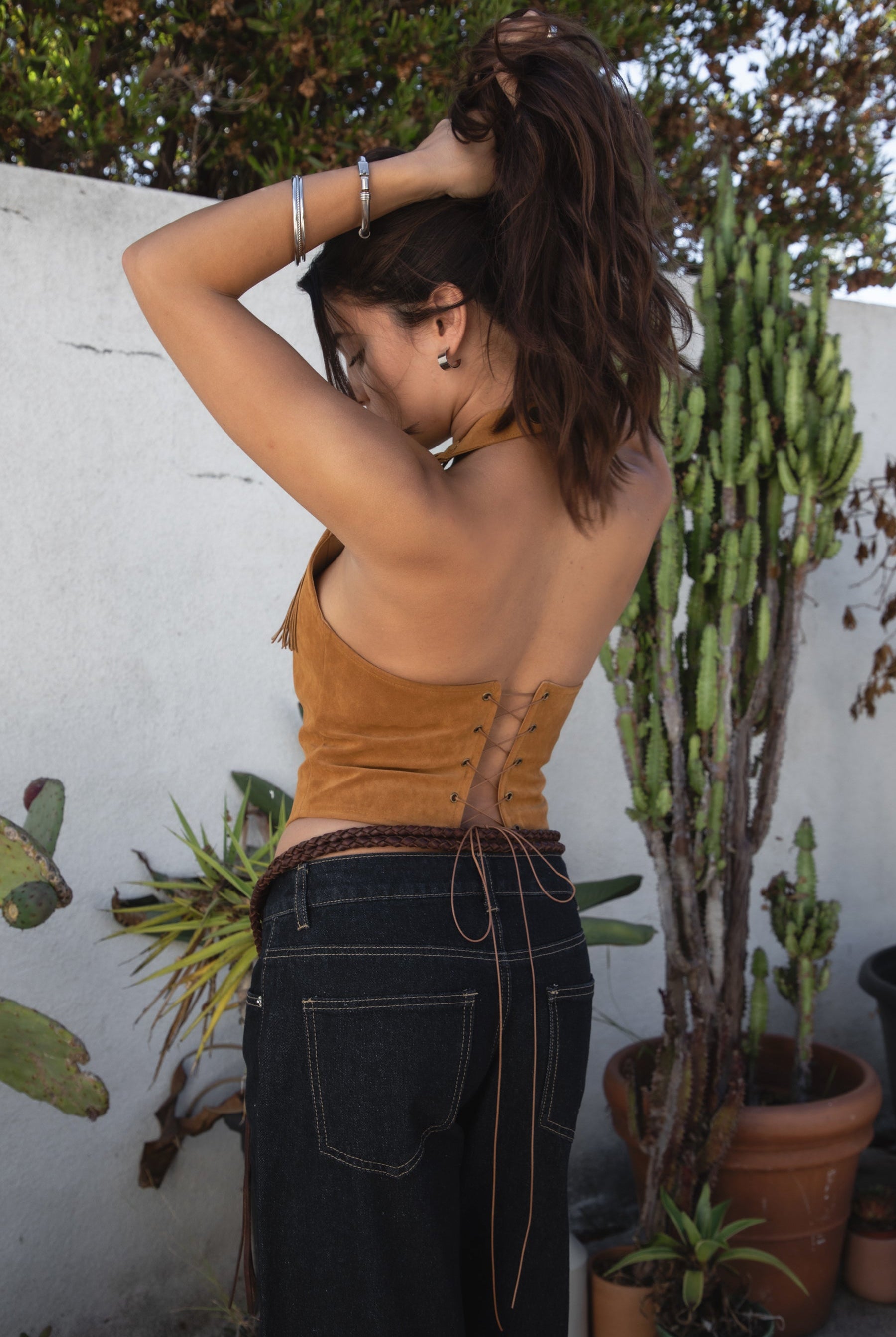 Woman wearing the TIC TOC TA31474 JOSIE tan faux suede fringe halter top featuring layered fringe neckline, lace trim tie detail, and open back with lace-up closure and dark jeans against a white wall and cacti in the background