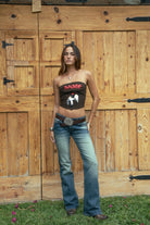 Woman wearing the TIC TOC TA31571 RODEA strapless western bustier top featuring sequin RODEO lettering, horse patch, and lace-up back closure and jeans against a wooden background