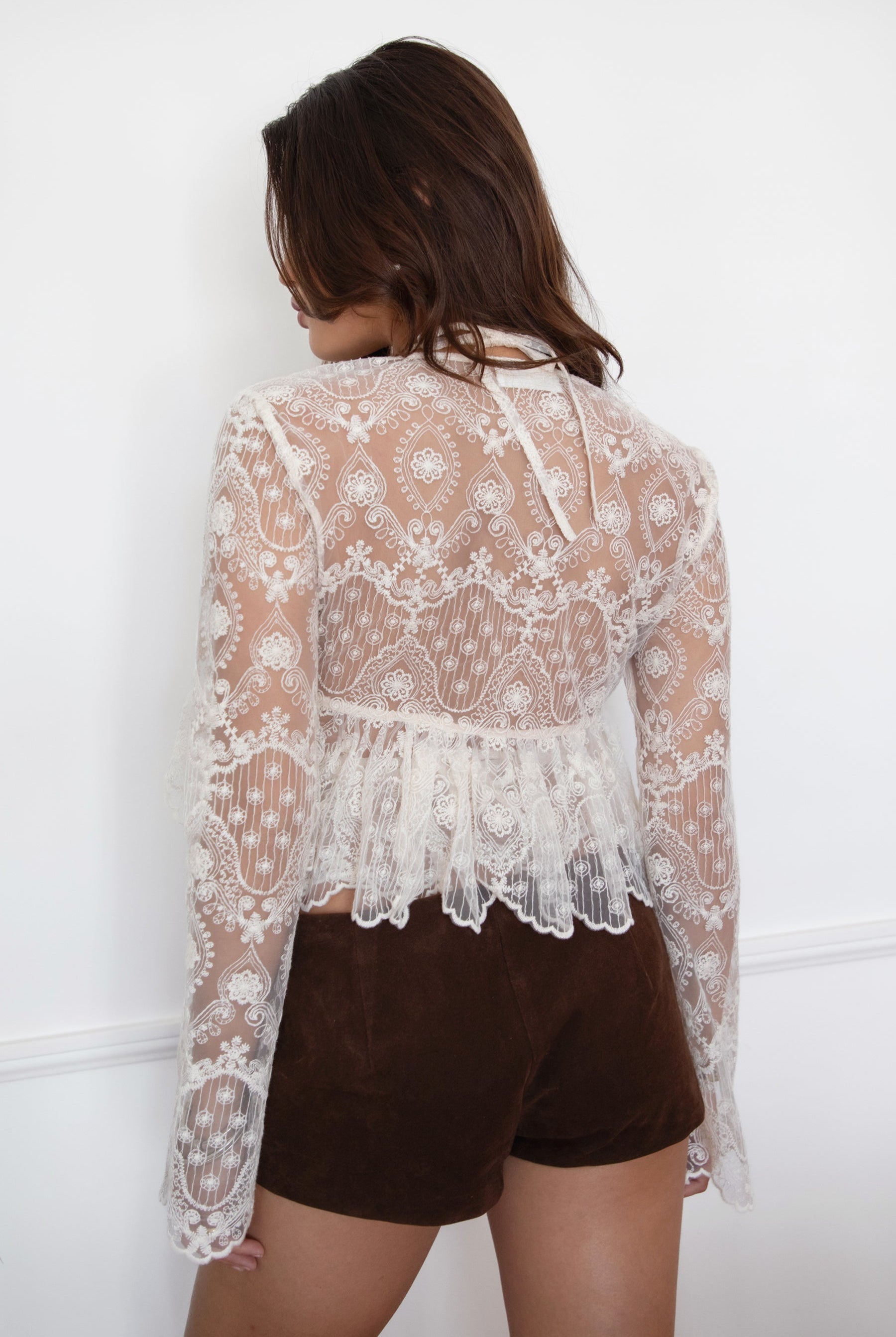 Model wearing the TIC TOC TA31608 LAREDA cream crochet embroidered long sleeve top featuring a scalloped V-neckline, tie front detail, and flowy bell sleeves with brown mini shorts.