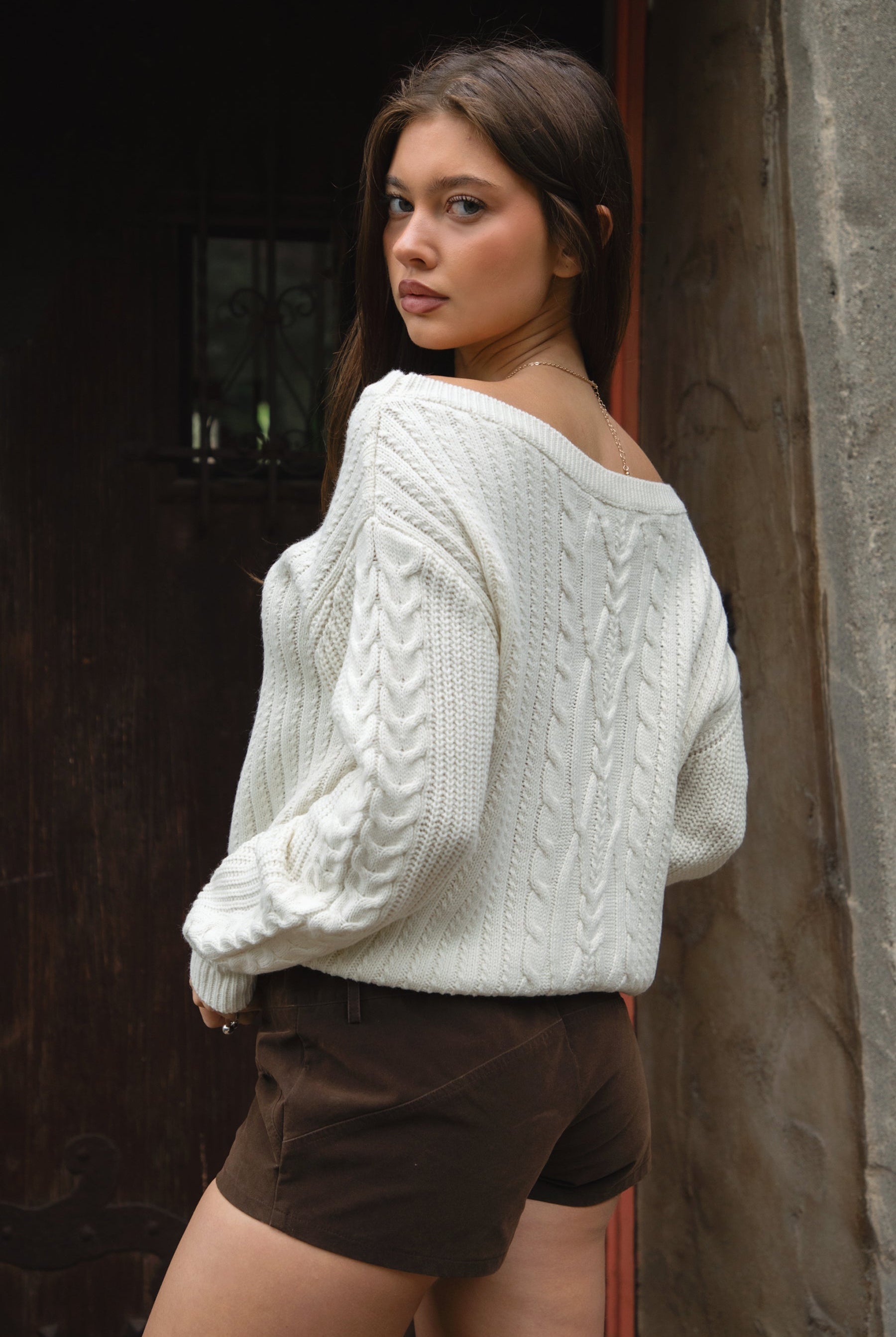 Woman wearing TIC TOC TA31803-ISARA cream oversized cable knit sweater with off-the-shoulder ribbed neckline and relaxed fit and brown shorts against a dark background.