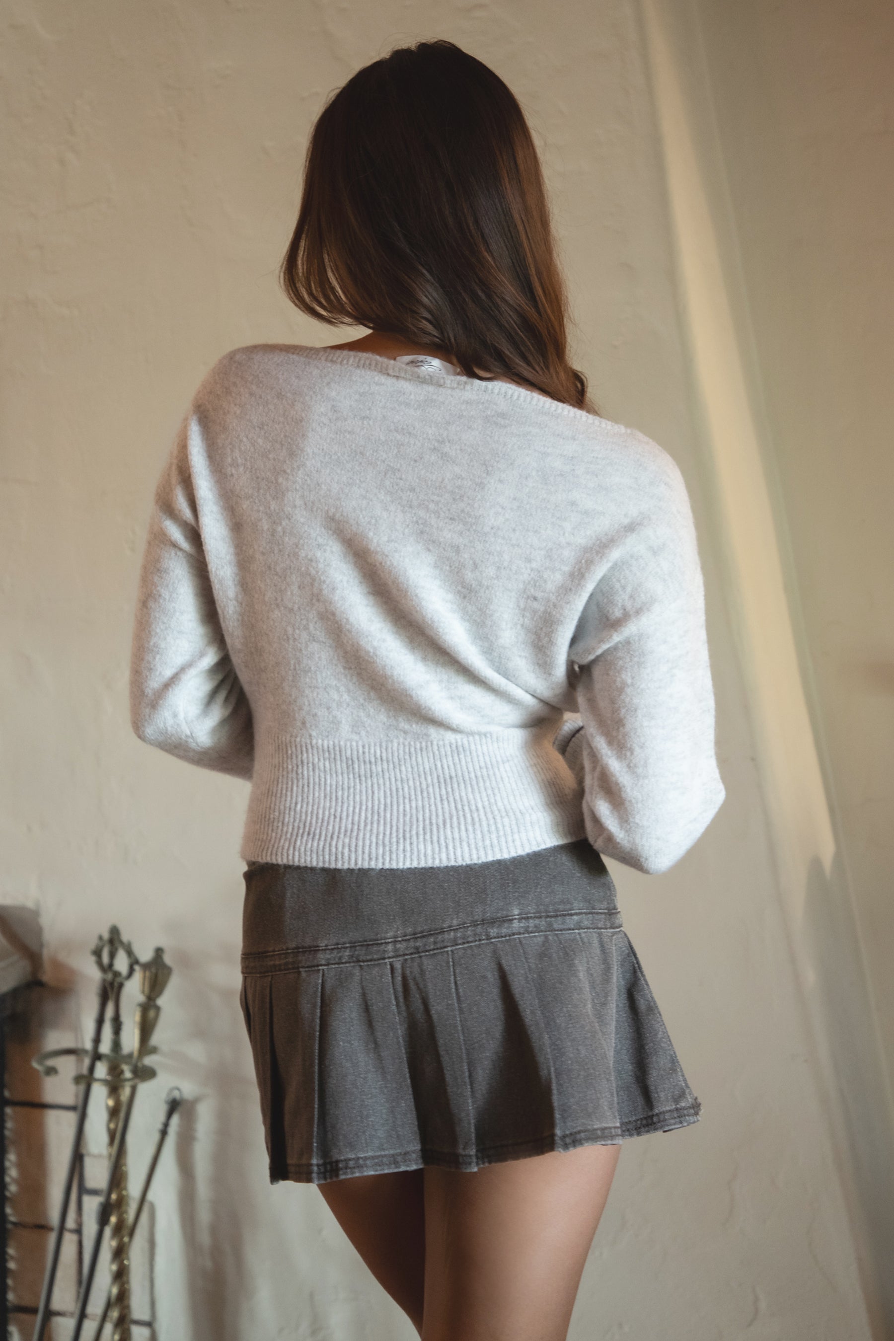 Woman wearing TIC TOC TA31843 AURELLE heather grey off the shoulder knit sweater top with dropped shoulders and relaxed long sleeves paired with a pleated brown mini skirt in an indoor setting.