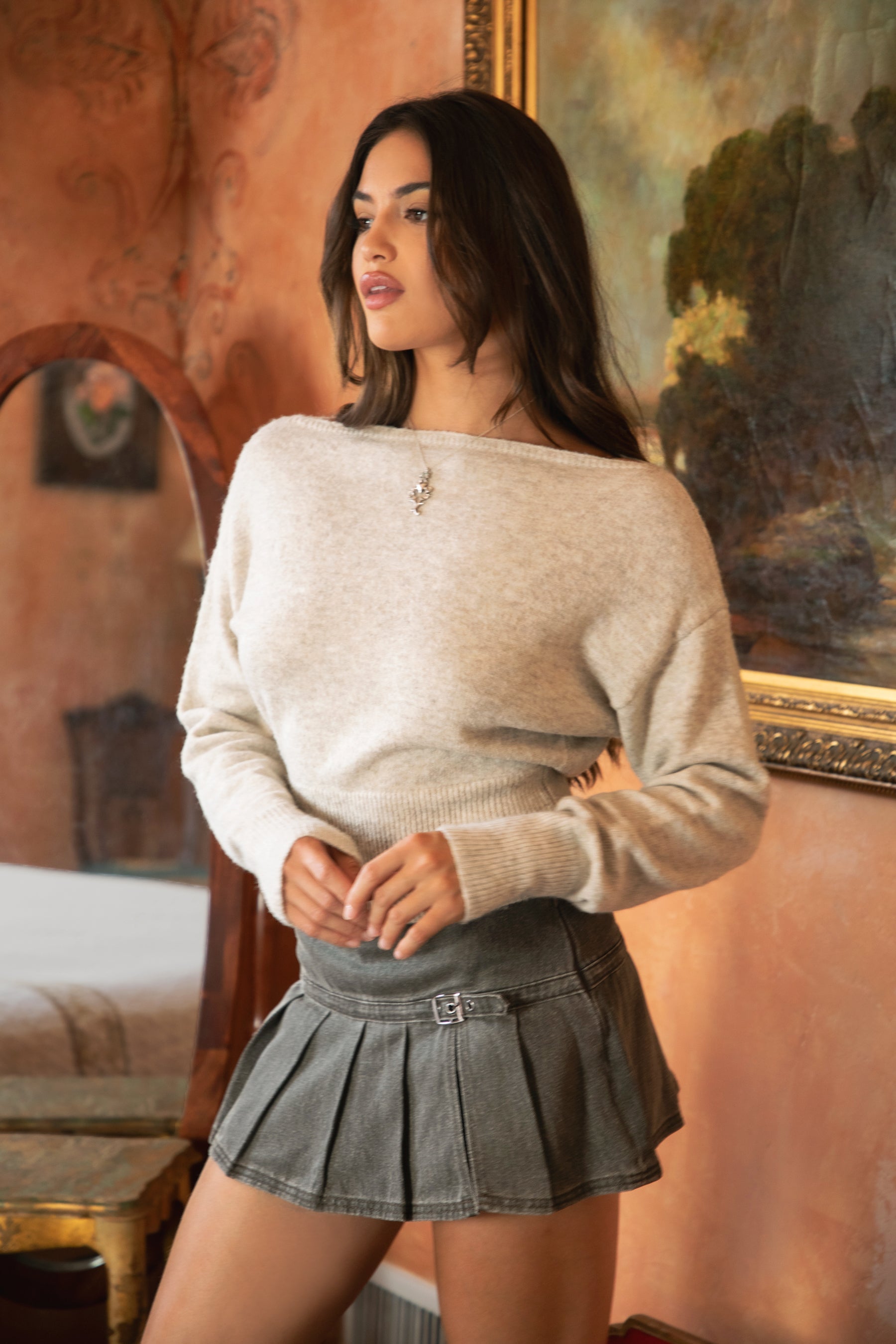 Woman wearing TIC TOC TA31843 AURELLE oatmeal off the shoulder knit sweater top with dropped shoulders and relaxed long sleeves paired with a pleated brown mini skirt in an indoor setting.